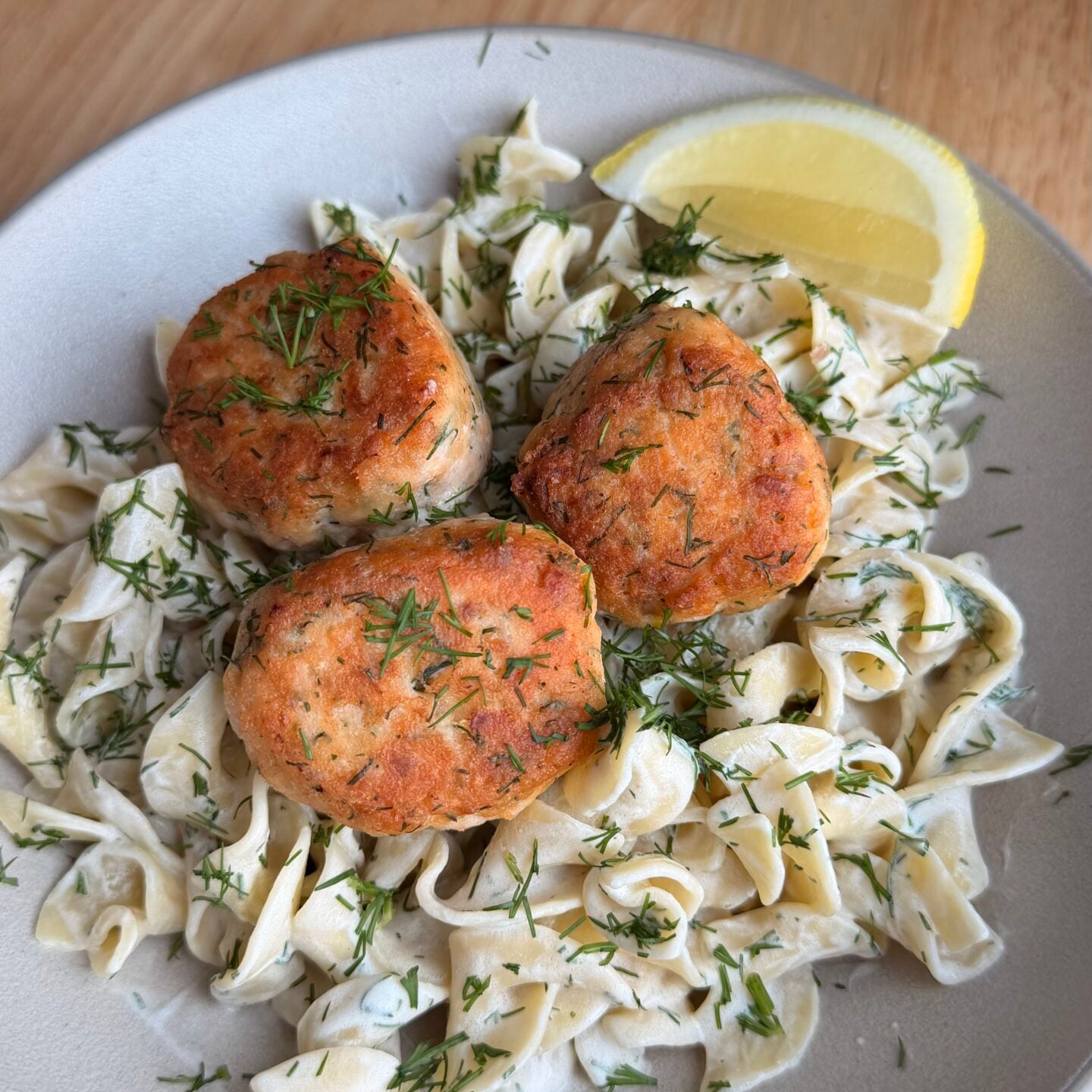 Three golden brown fish cakes on a bed of pasta with creamy sauce, sprinkled with herbs, and served with two lemon wedges on a gray plate.