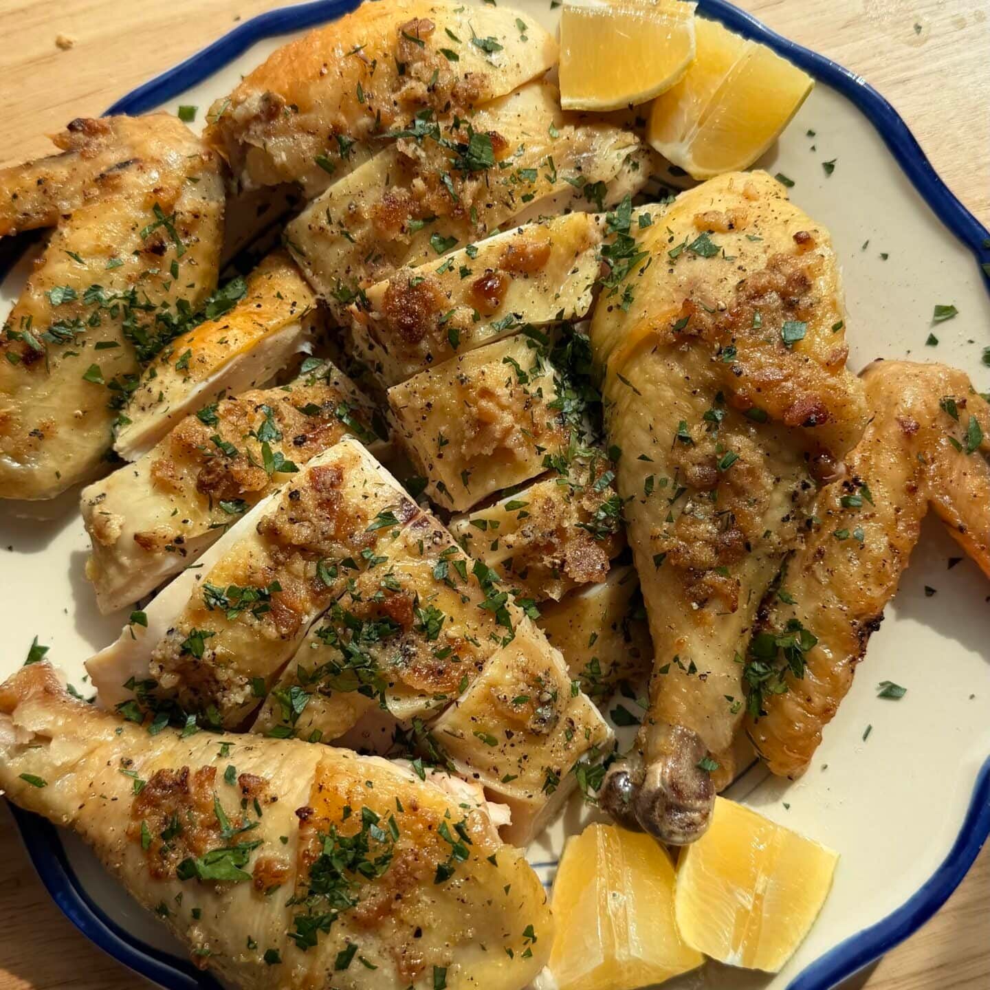 A plate of roasted chicken pieces garnished with herbs, accompanied by lemon wedges, on a wooden surface in natural sunlight.