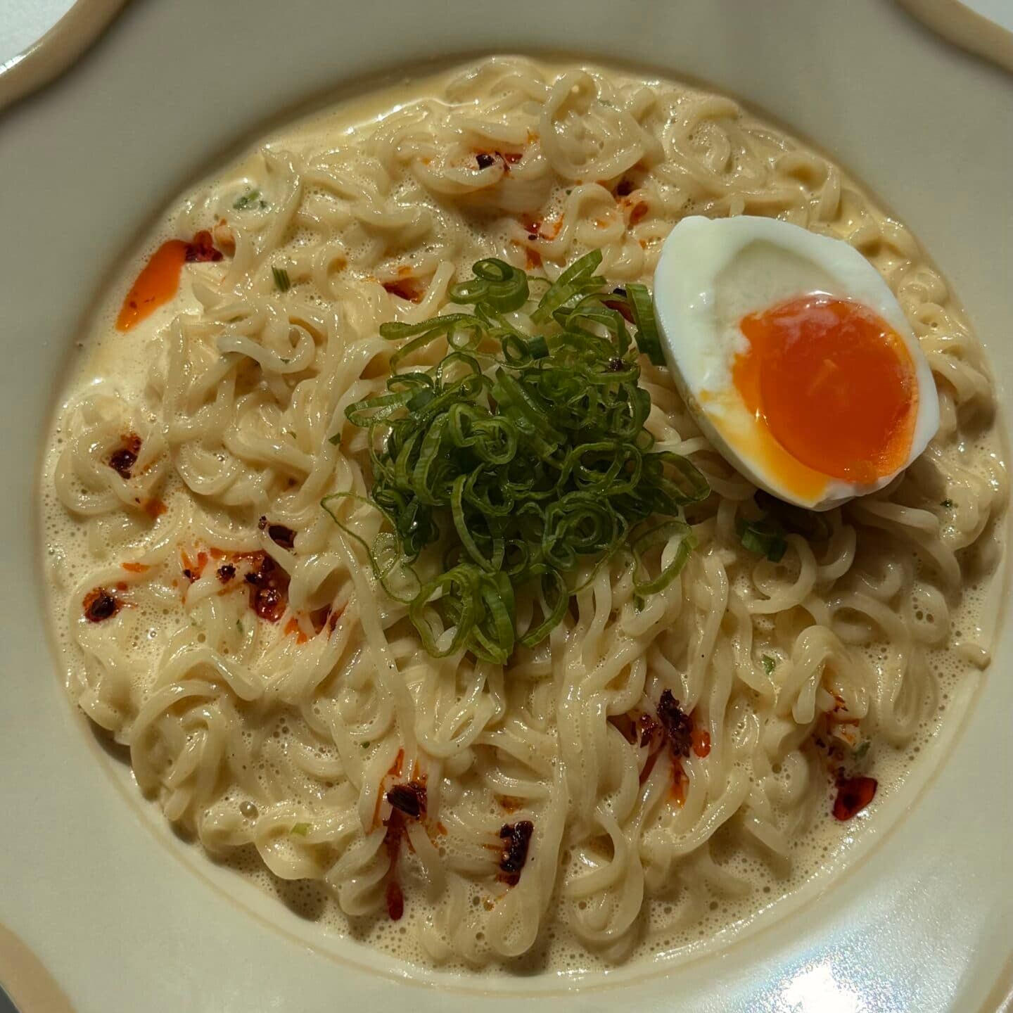 Bowl of ramen noodles in creamy broth, topped with sliced green onions, chili flakes, and half of a soft-boiled egg.