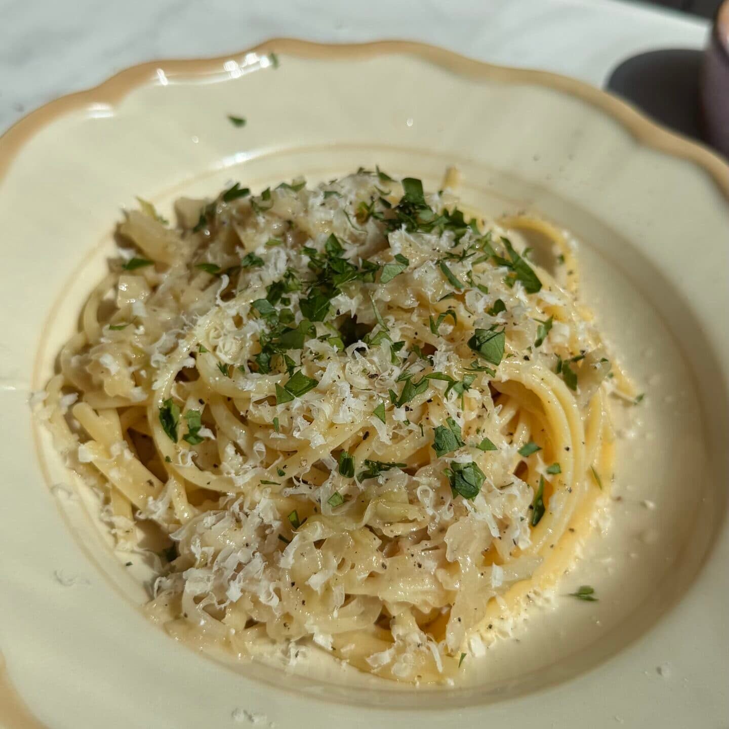 A plate of spaghetti topped with grated cheese and chopped herbs, served on a cream-colored dish on a white marble surface.