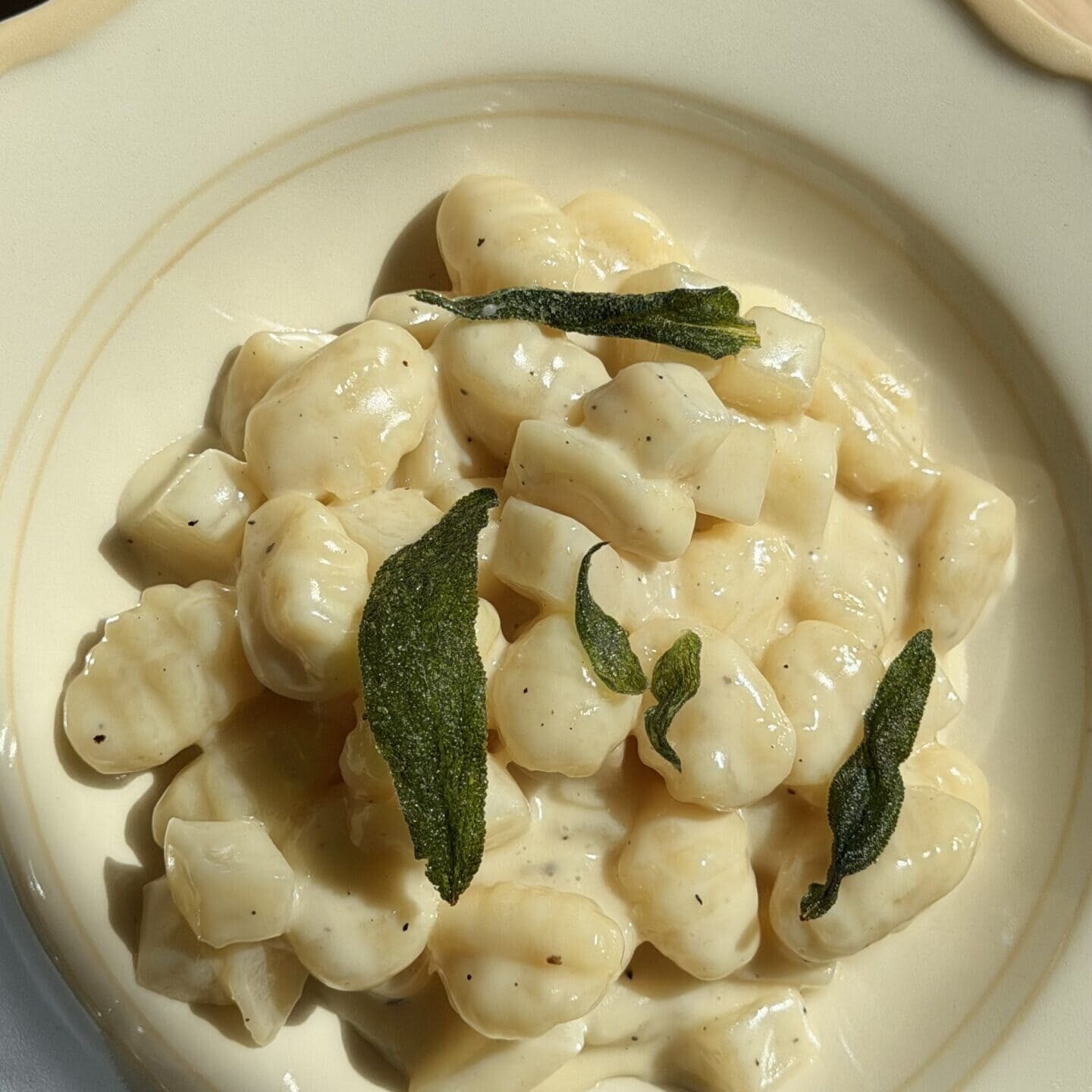 A plate of gnocchi in a creamy white sauce, garnished with crispy sage leaves, served in a cream-colored bowl.