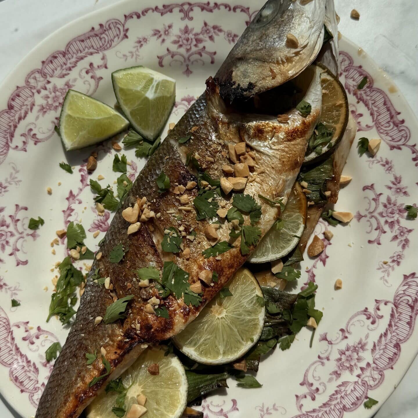 A whole roasted fish garnished with chopped herbs and nuts, served on a plate with lime slices and lime wedges.