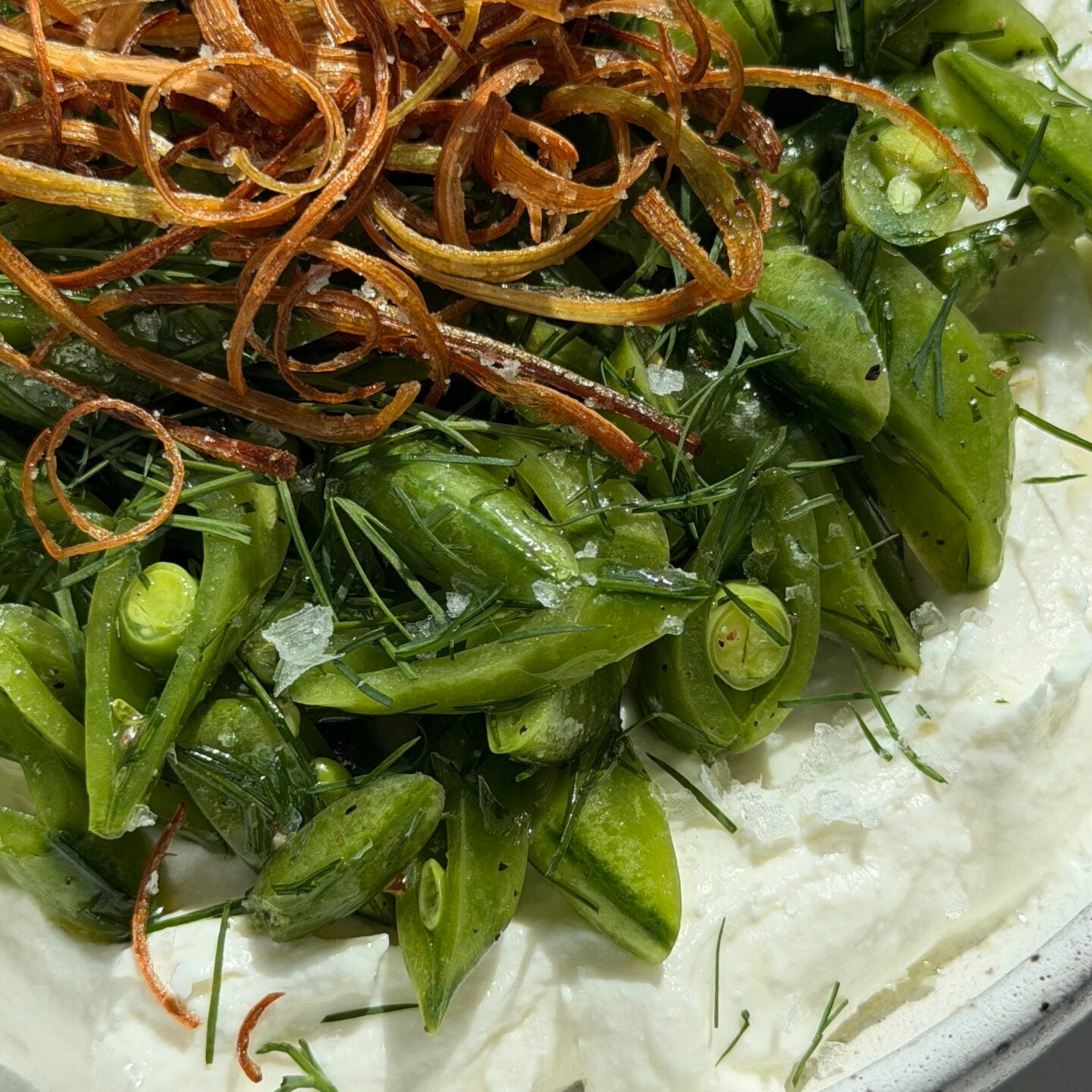 A plate of whipped ricotta topped with sliced snap peas, fresh herbs, and crispy fried shallots.