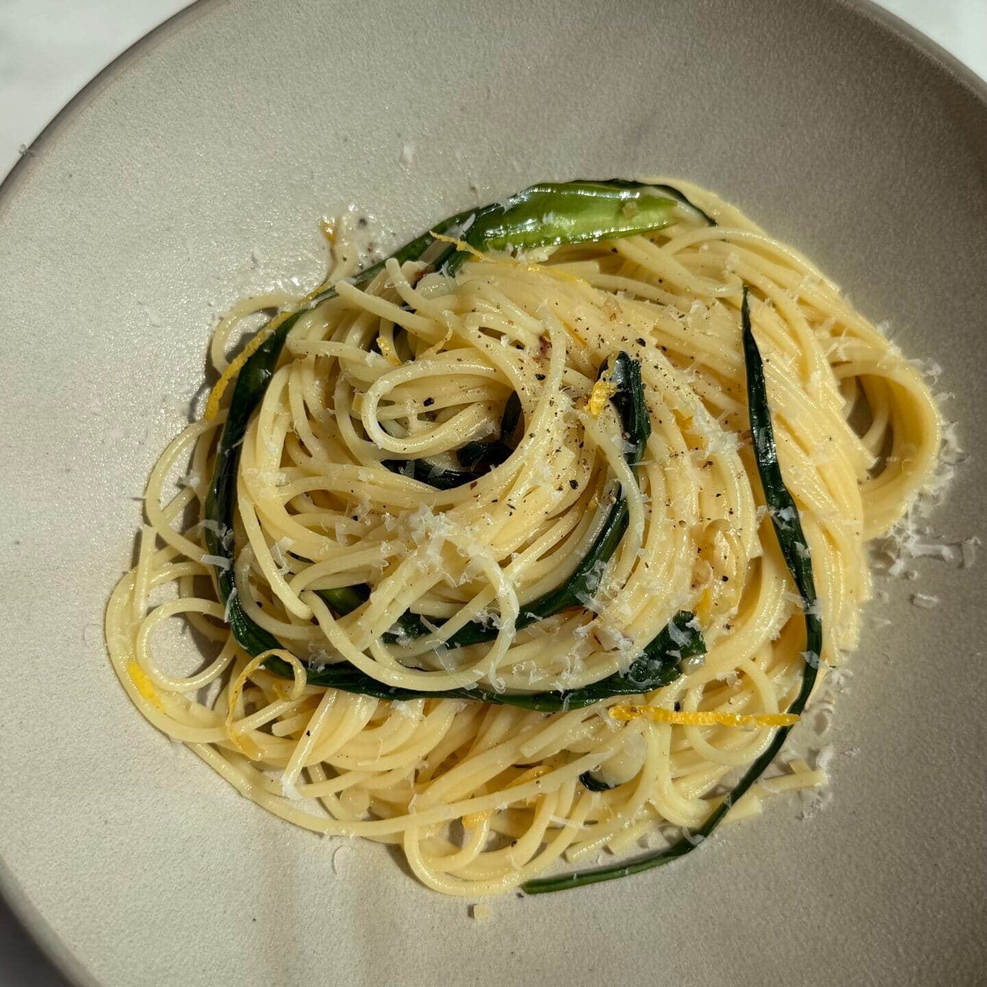 A plate of spaghetti topped with grated cheese and green vegetables, served on a light-colored dish against a marble background.