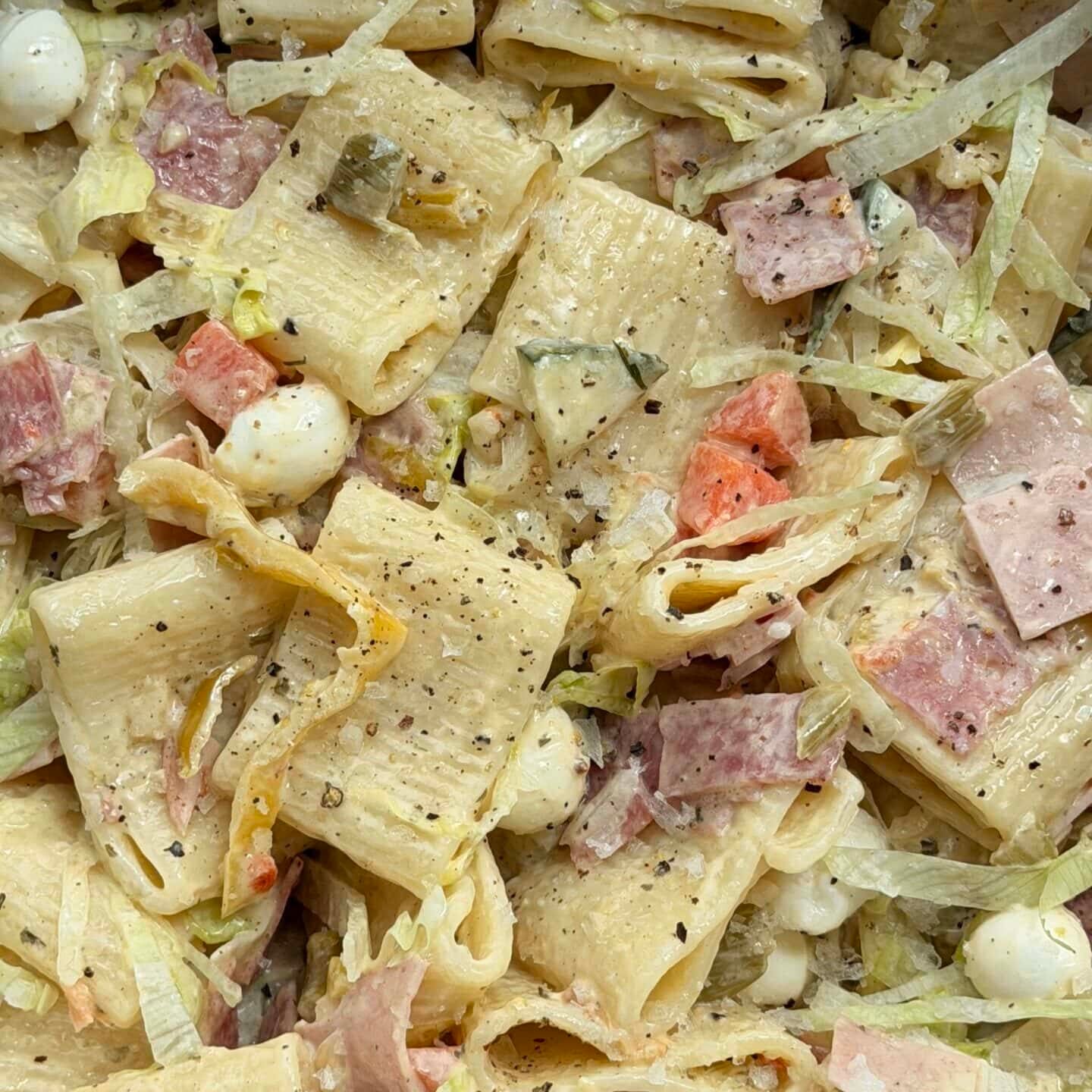A close-up of pasta salad with rigatoni, diced deli meats, mozzarella balls, lettuce, pickles, tomatoes, and black pepper in a creamy dressing.