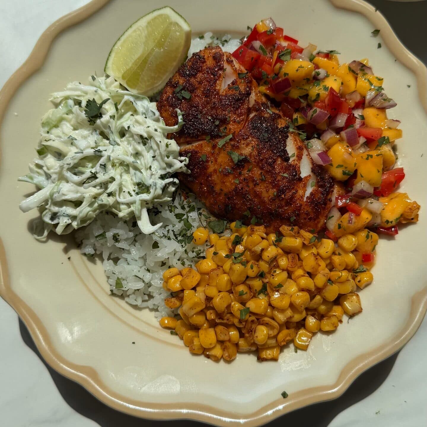 A plate with blackened fish, mango salsa, grilled corn, shredded cabbage, a lime wedge, and rice on a beige dish.