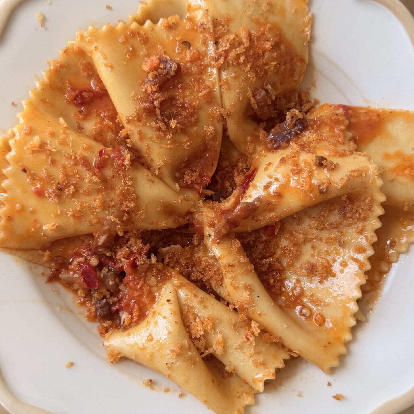 A white plate with wide, square pasta pieces topped with a chunky red sauce and sprinkled with grated cheese or breadcrumbs.
