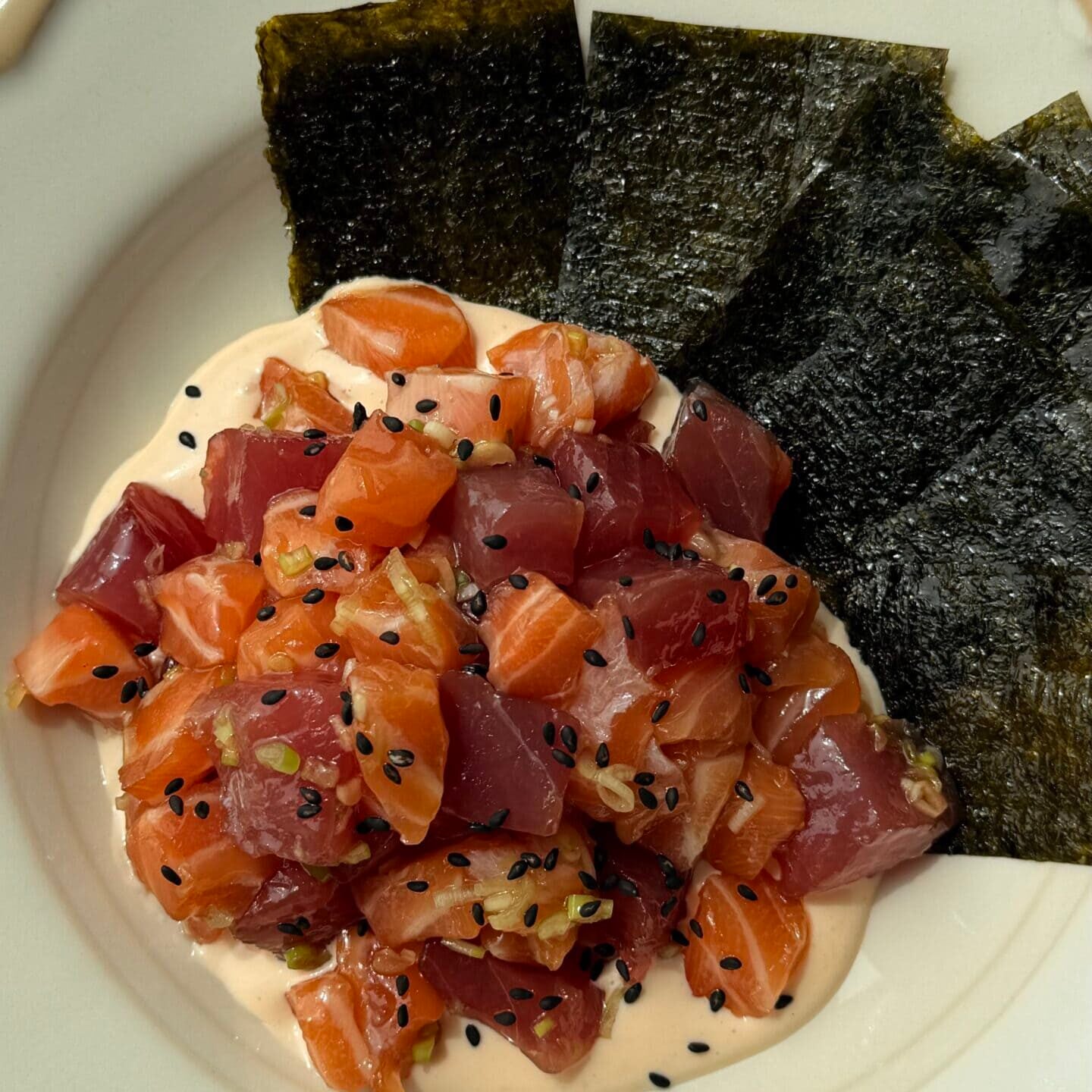 A plate with diced raw salmon and tuna on a creamy sauce, topped with black sesame seeds, served with sheets of seaweed on the side.