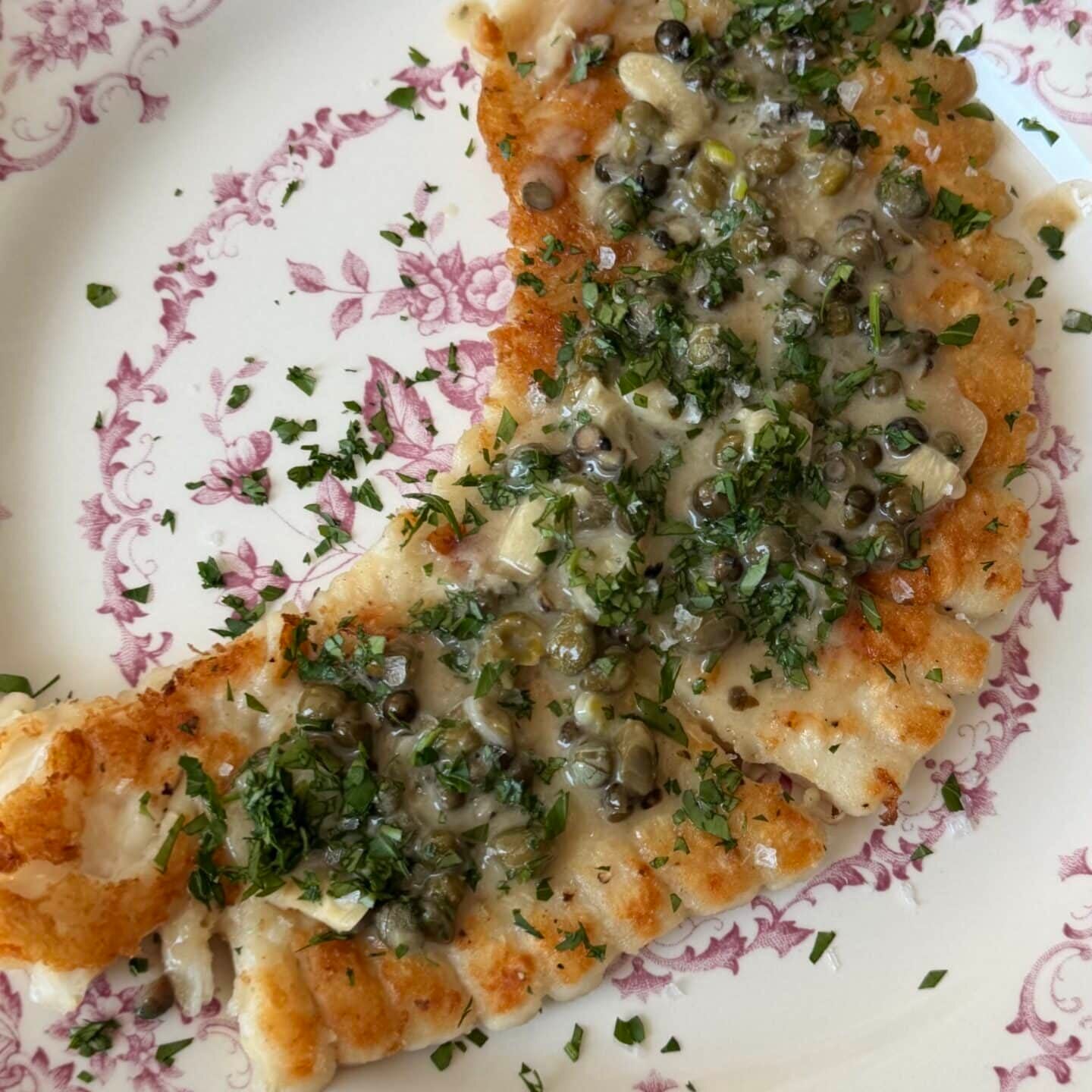 A browned, crescent-shaped piece of fish topped with capers and chopped herbs on a white and pink decorative plate.