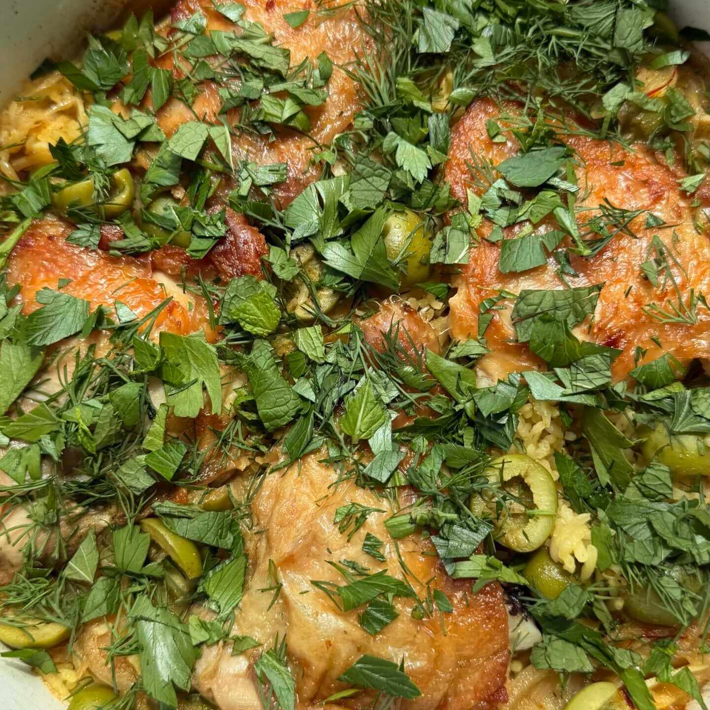 A pot of cooked chicken thighs topped with chopped fresh herbs and sliced green olives.