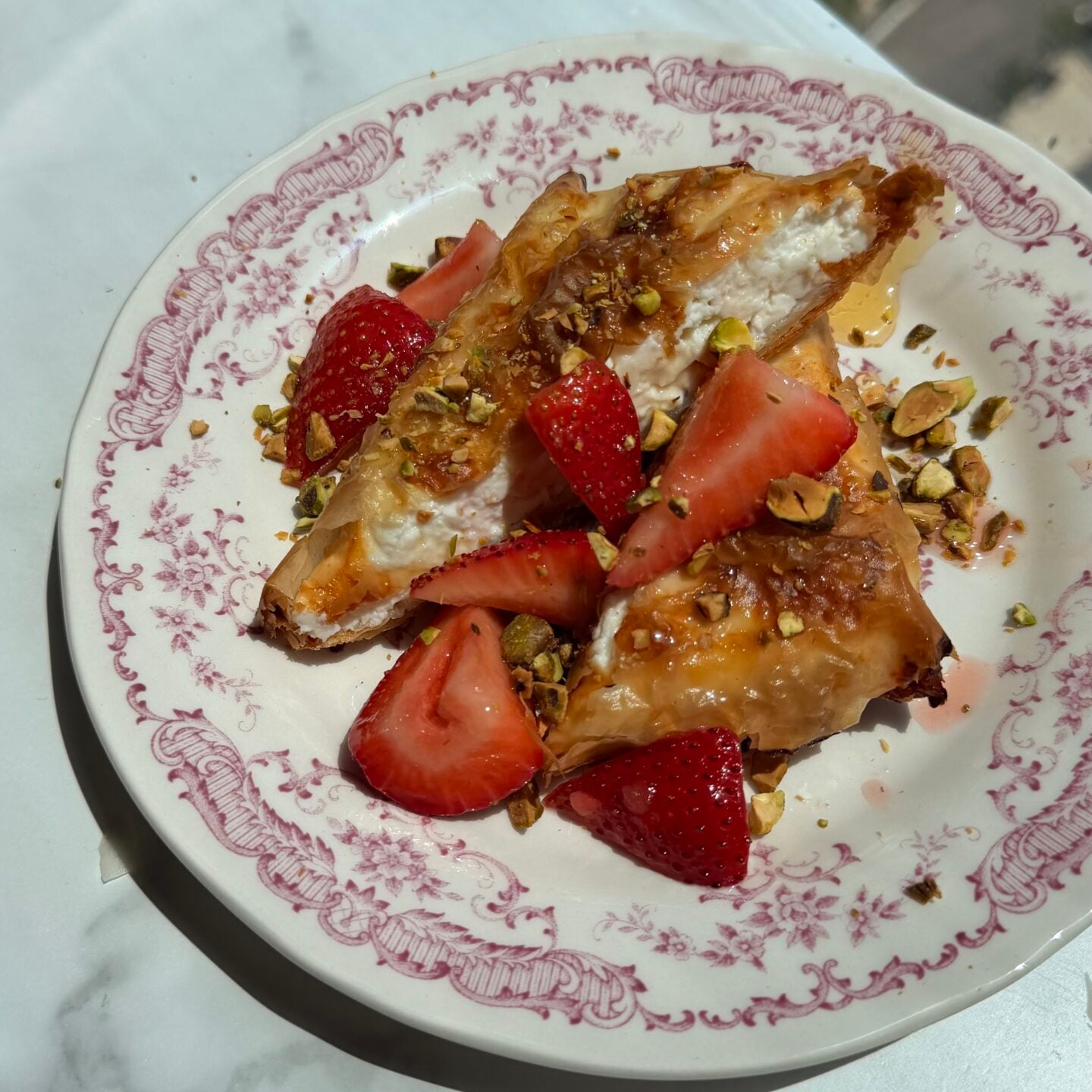 A pastry topped with fresh strawberries and chopped pistachios is served on a decorative plate, placed on a white surface near a window.