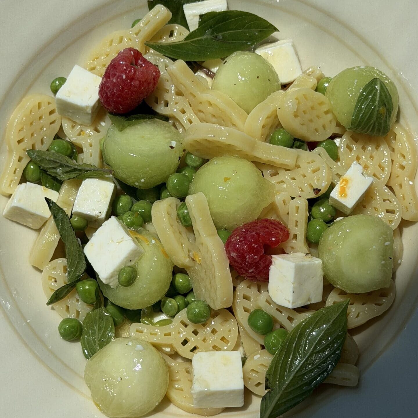 A bowl of wheel-shaped pasta with green peas, melon balls, feta cheese cubes, fresh basil leaves, and raspberries on a cream-colored plate.