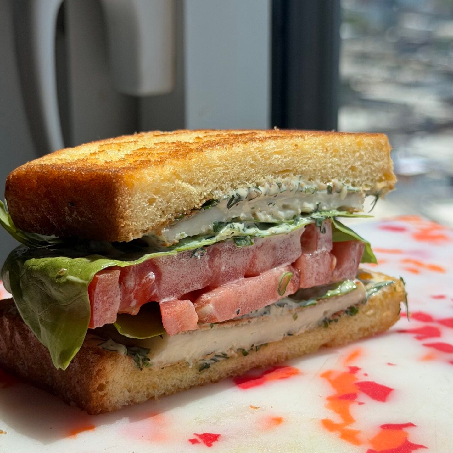 A close-up of a sandwich with lettuce, tomato, and herb spread on toasted bread, placed on a cutting board near a window with a city view in the background.