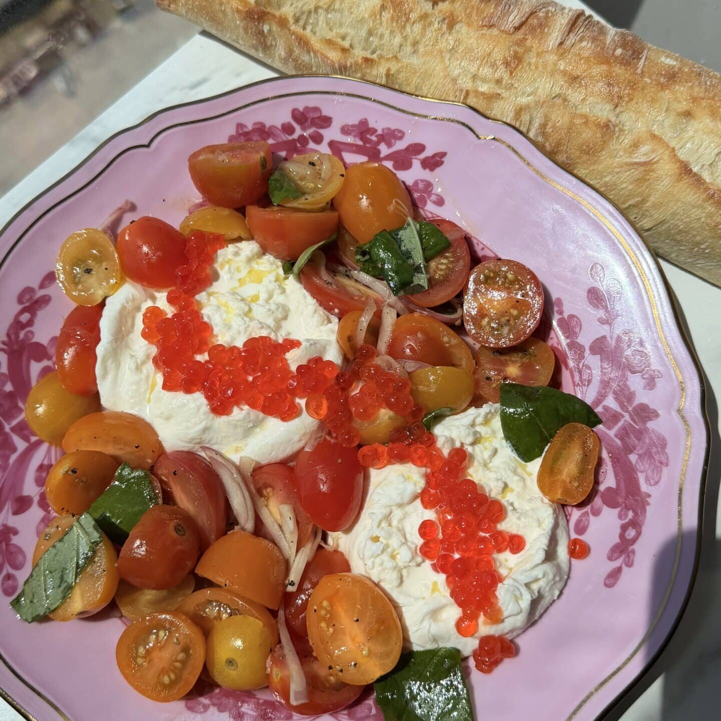 A plate of burrata cheese with cherry tomatoes, red roe, basil, and onions, served with a baguette, placed by a window in sunlight.