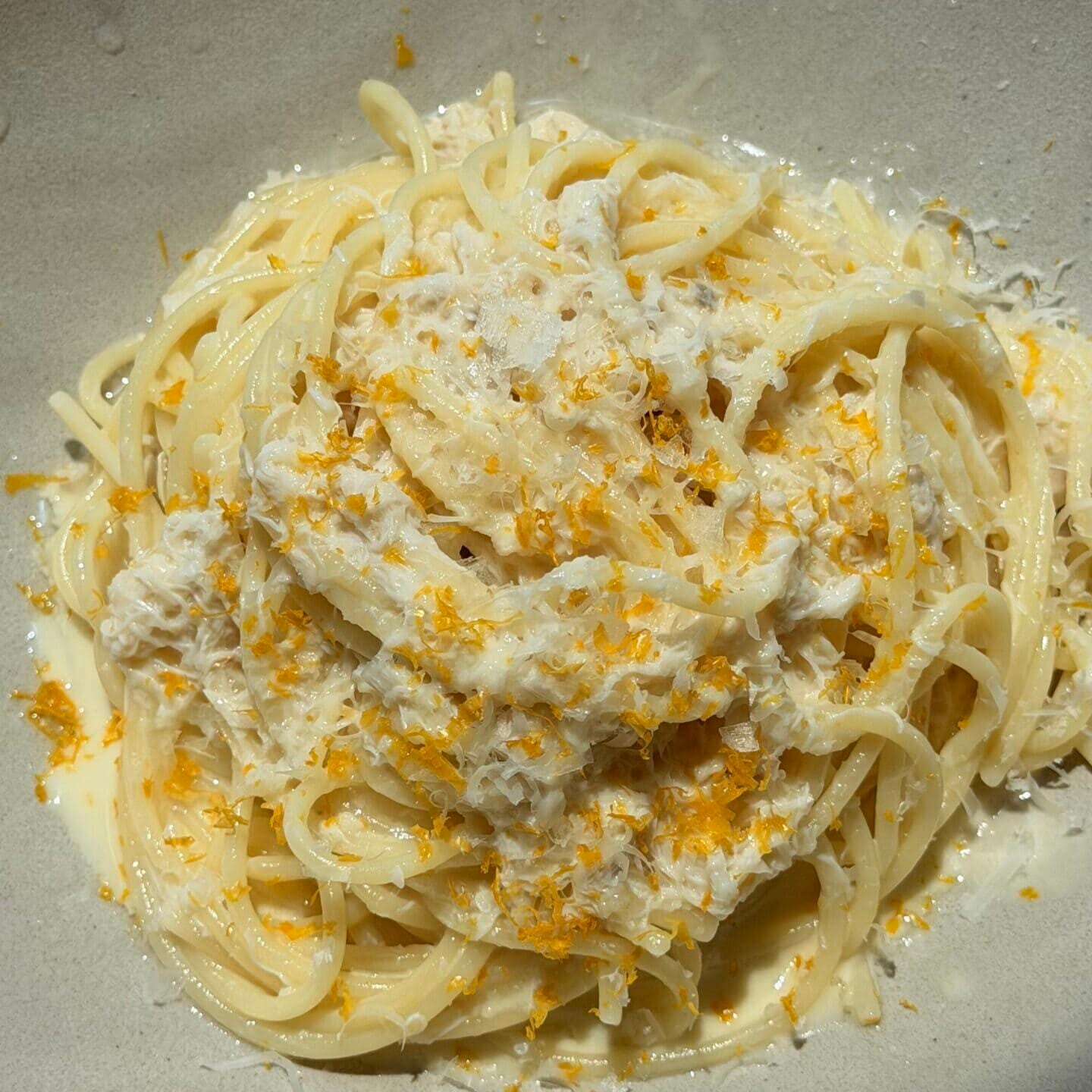 A bowl of spaghetti topped with a creamy white sauce, shredded cheese, and orange zest on a light-colored plate.