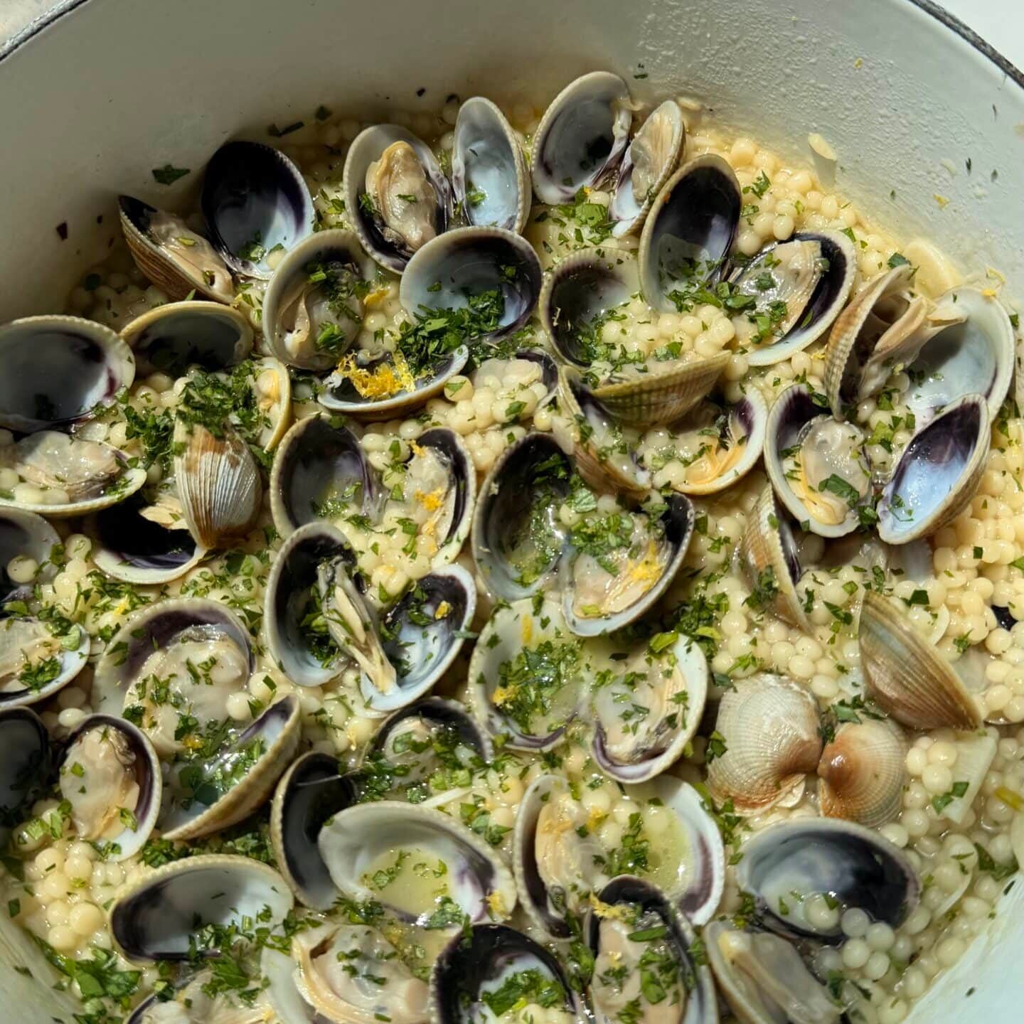 A white pot filled with cooked clams over pearl couscous, garnished with chopped herbs and lemon zest, sits by a sunlit window.