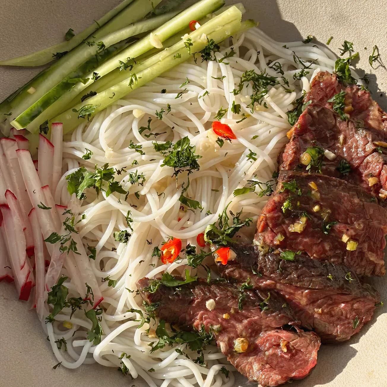 A bowl with rice noodles, sliced grilled beef, julienned cucumber and radish, garnished with chopped herbs and red chili.