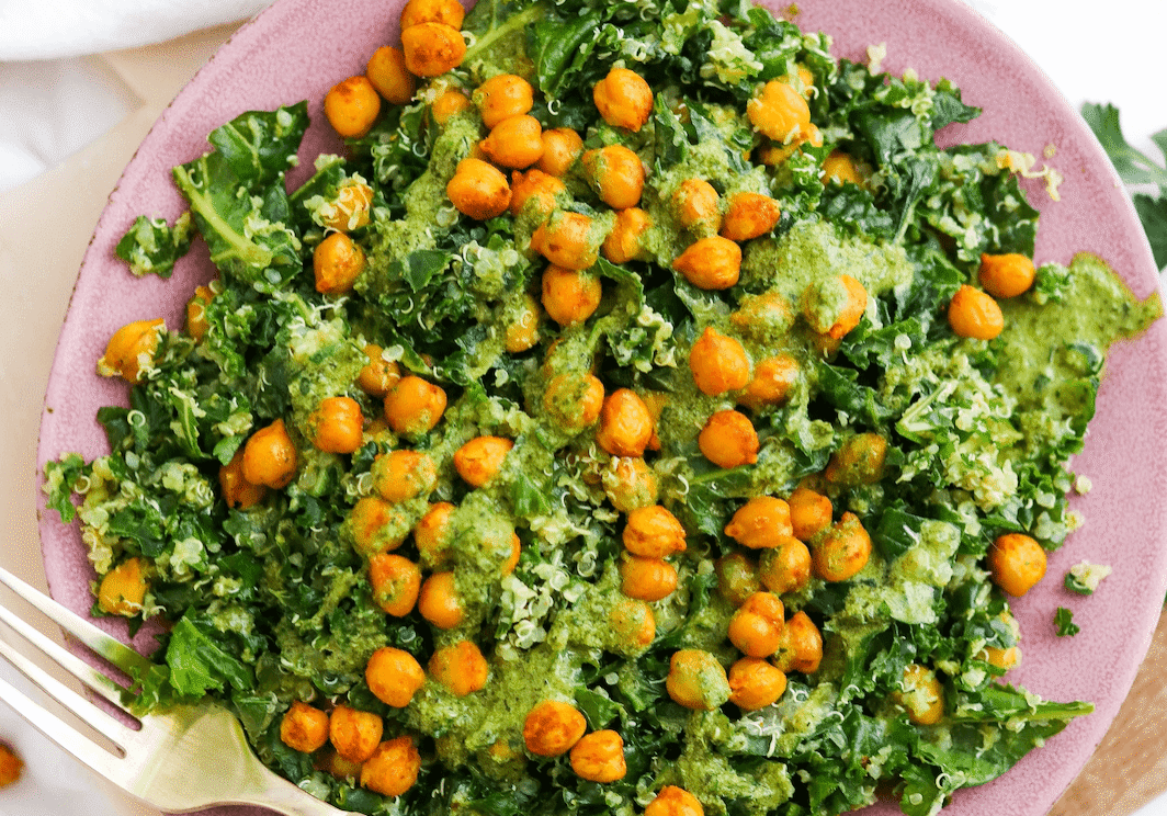 A plate of kale salad topped with roasted chickpeas and green dressing, with a fork placed on the side.