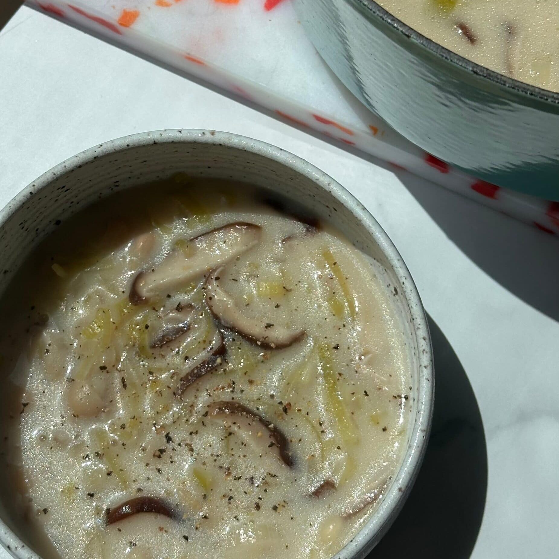 A bowl of creamy mushroom soup with visible mushroom slices and ground pepper, next to a pot containing more of the same soup.