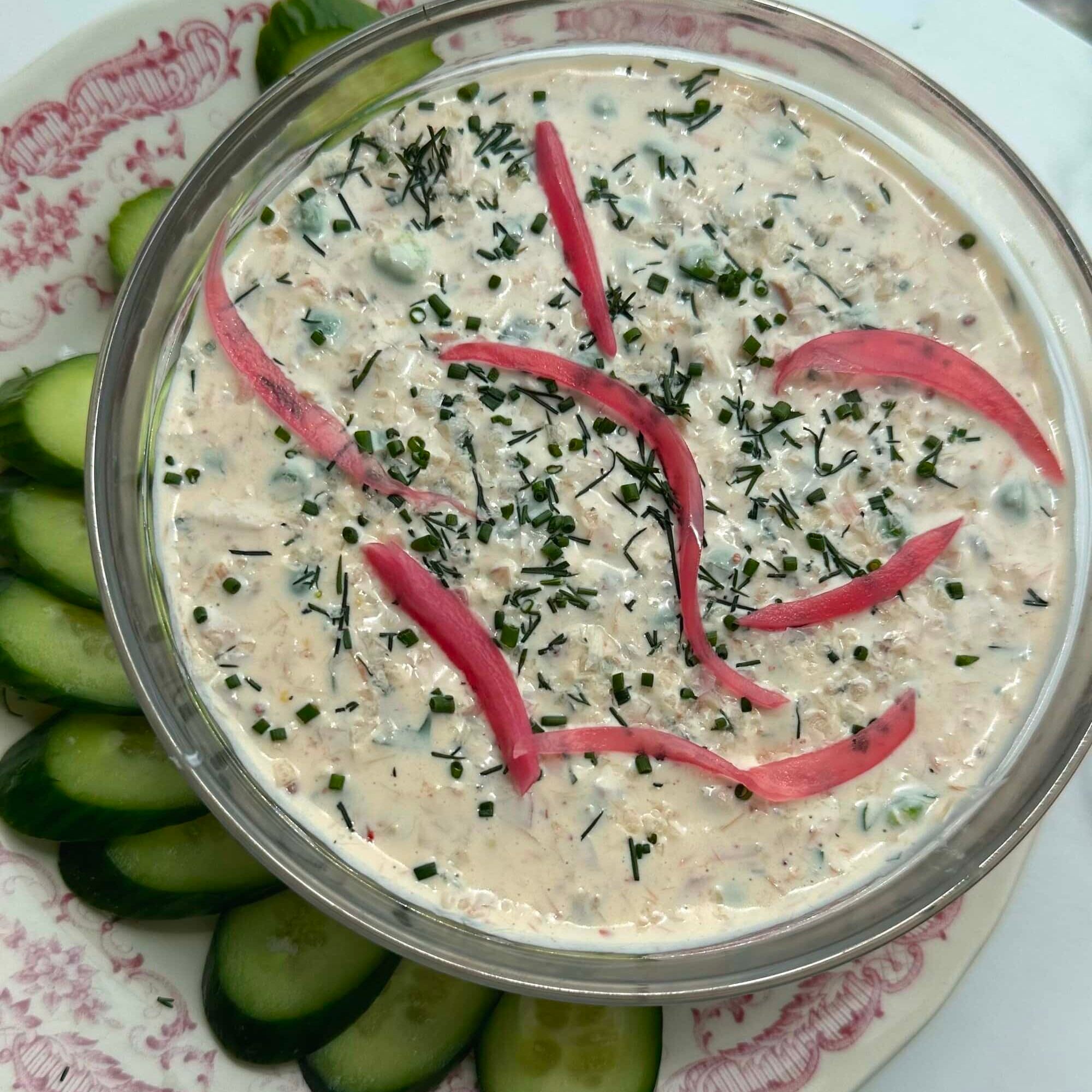 A bowl of creamy dip garnished with chopped herbs and pickled onions, surrounded by sliced cucumbers on a decorative plate.