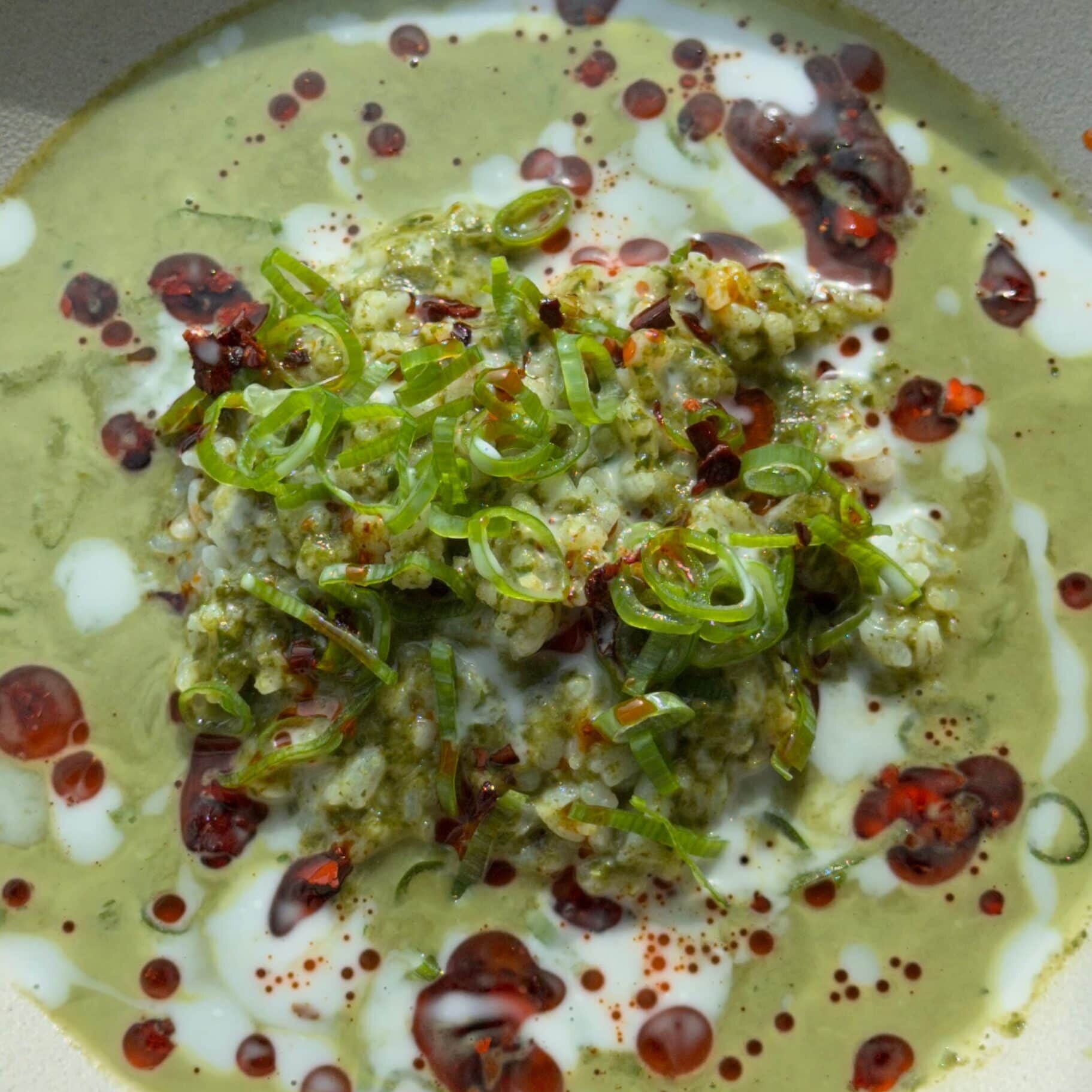 A bowl of green soup topped with a dollop of rice, sliced scallions, white cream, and red chili oil drizzles.
