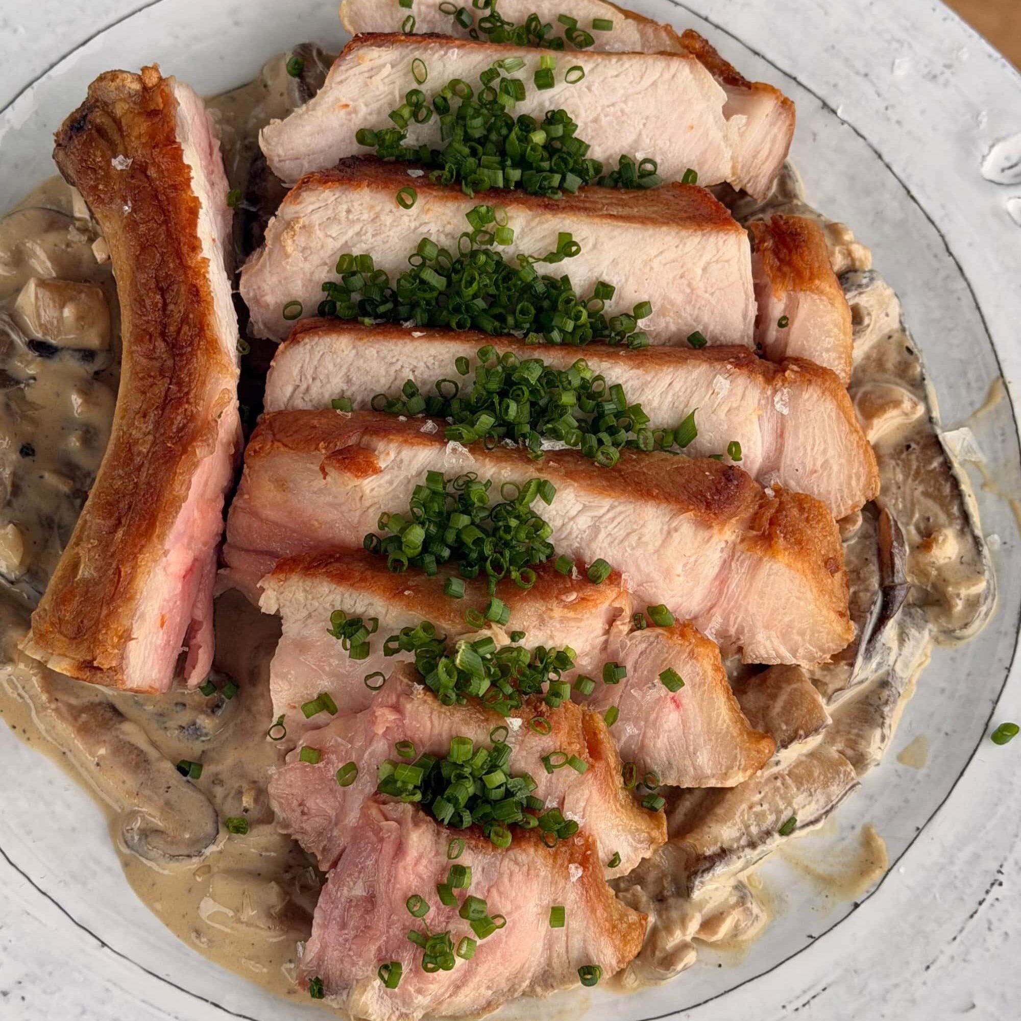 Sliced pork with a crispy edge served over creamy mushrooms, garnished with chopped chives on a white plate.