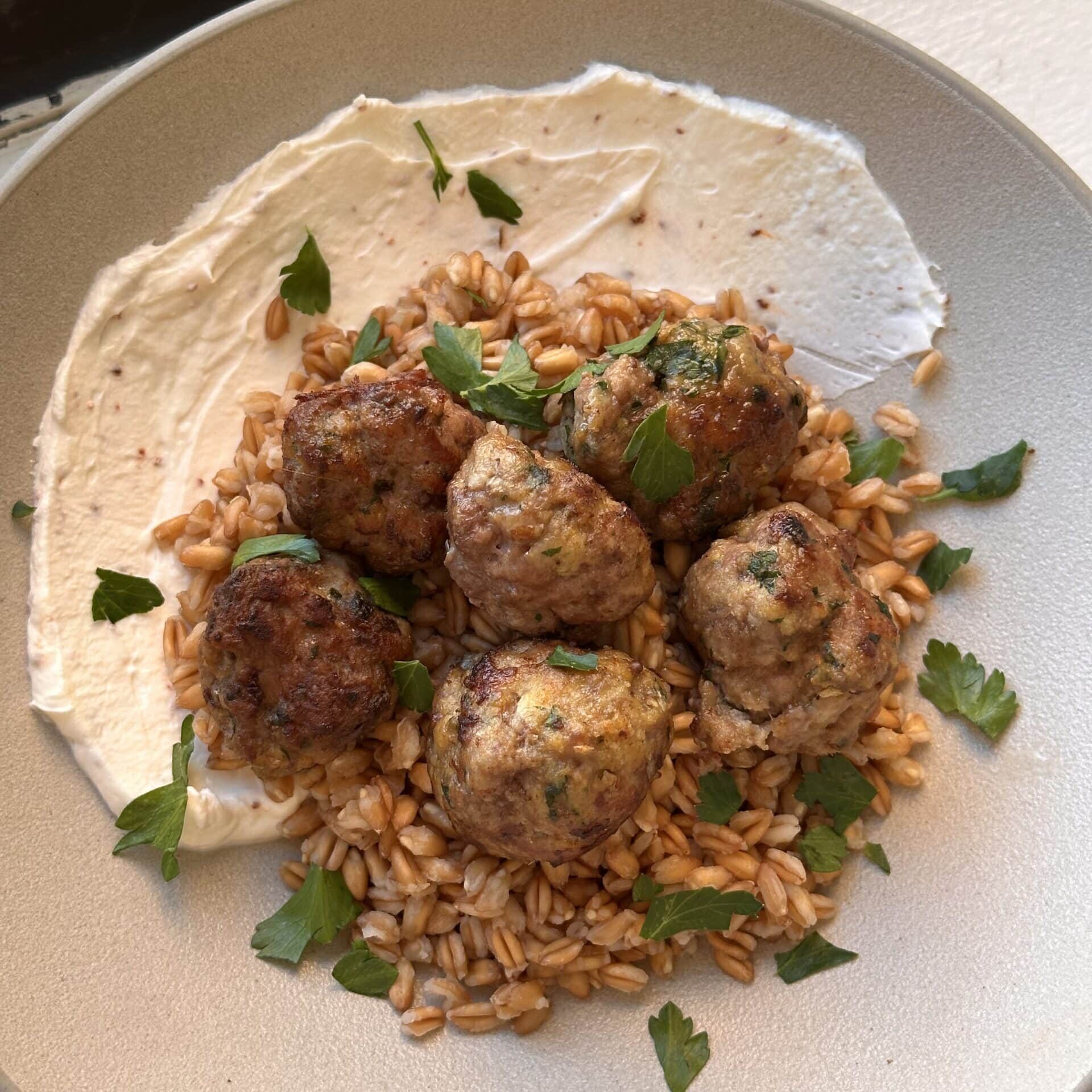 Five meatballs on a bed of grains, served with a smear of white sauce and garnished with fresh parsley on a beige plate.