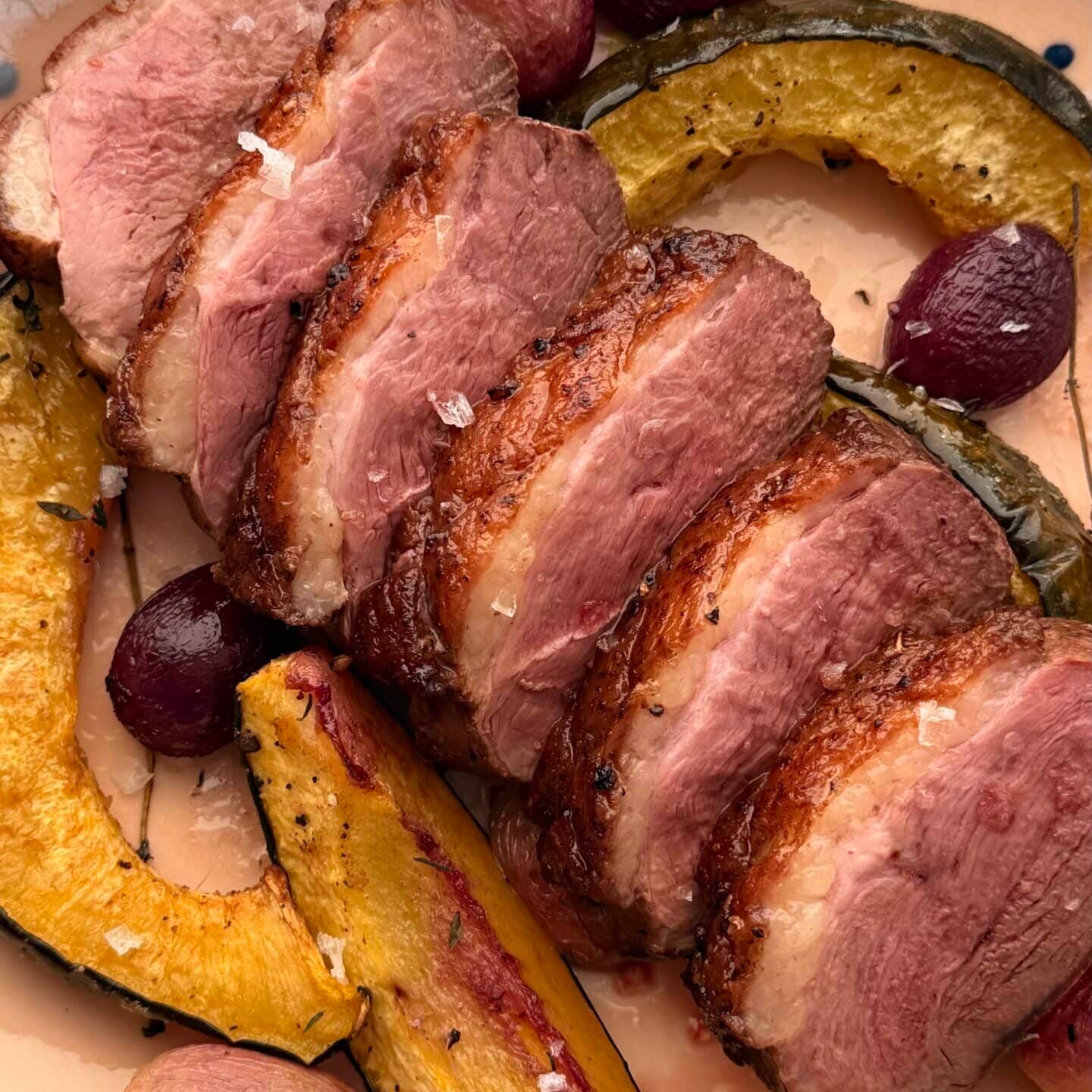 Sliced roasted duck breast served with roasted squash and red grapes on a speckled ceramic plate.