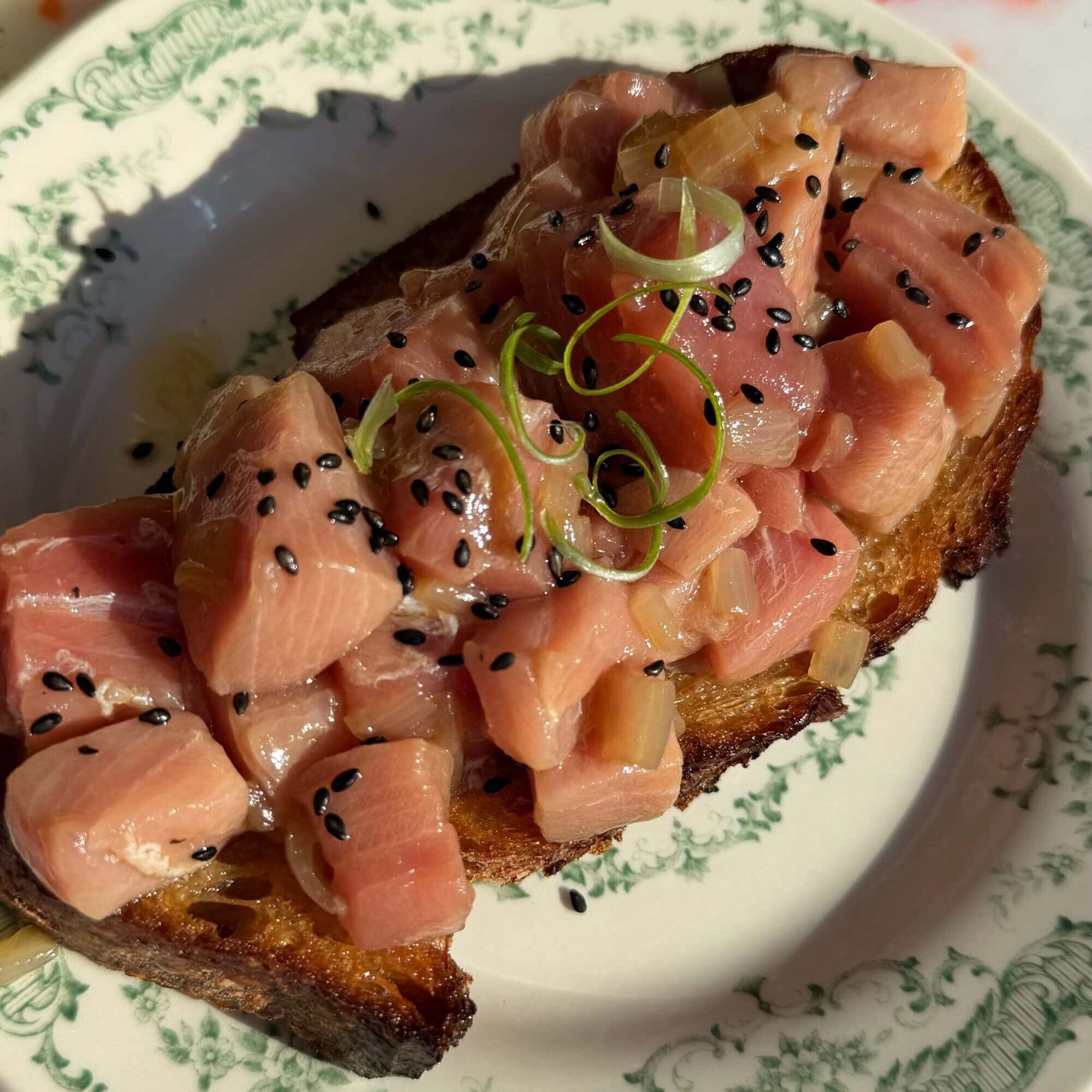 A slice of toasted bread topped with diced raw fish, garnished with black sesame seeds and thinly sliced green onions, served on a decorative plate.