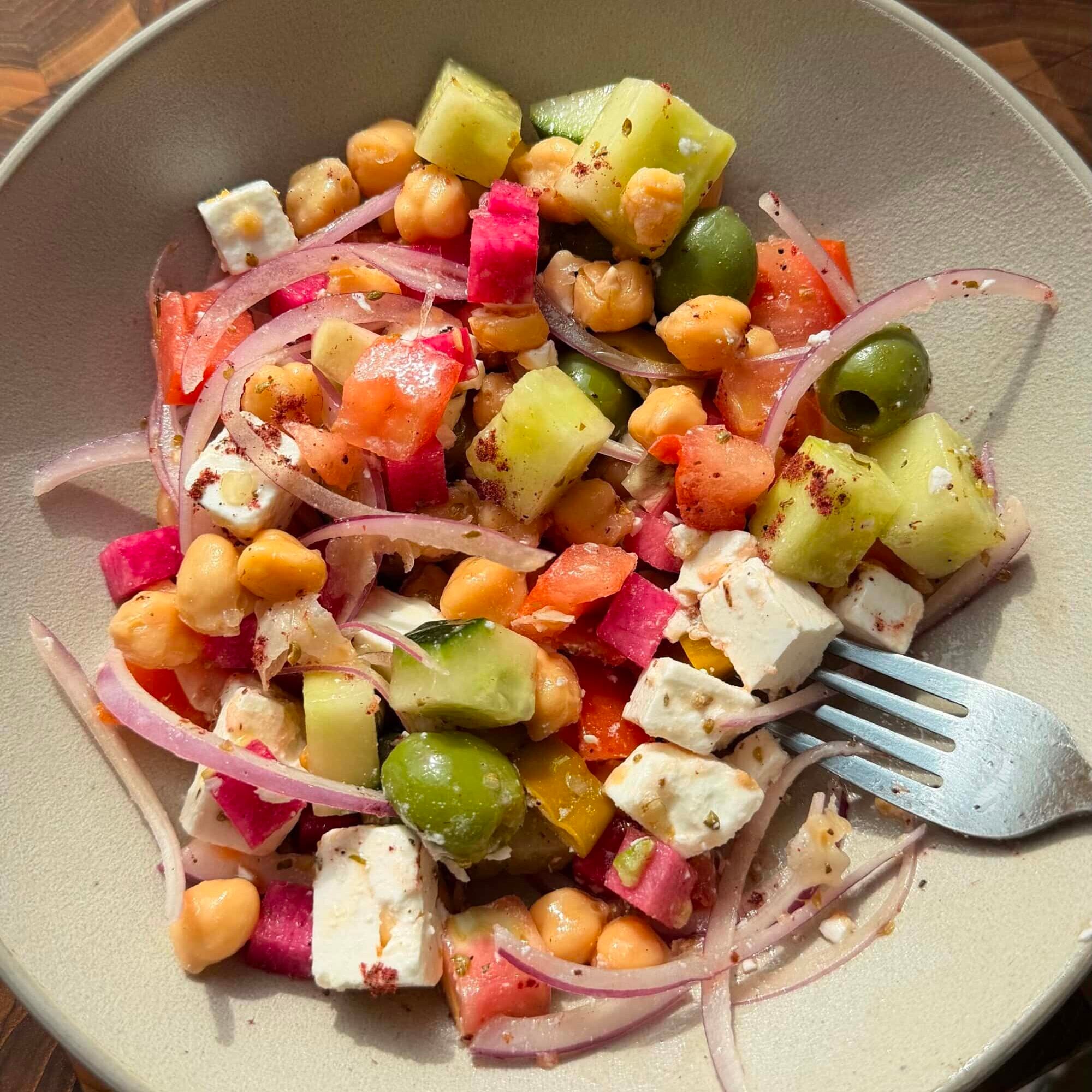 A bowl of salad with chickpeas, feta cheese, cucumber, green olives, diced tomatoes, red onion, and watermelon radish, with a fork on the side.