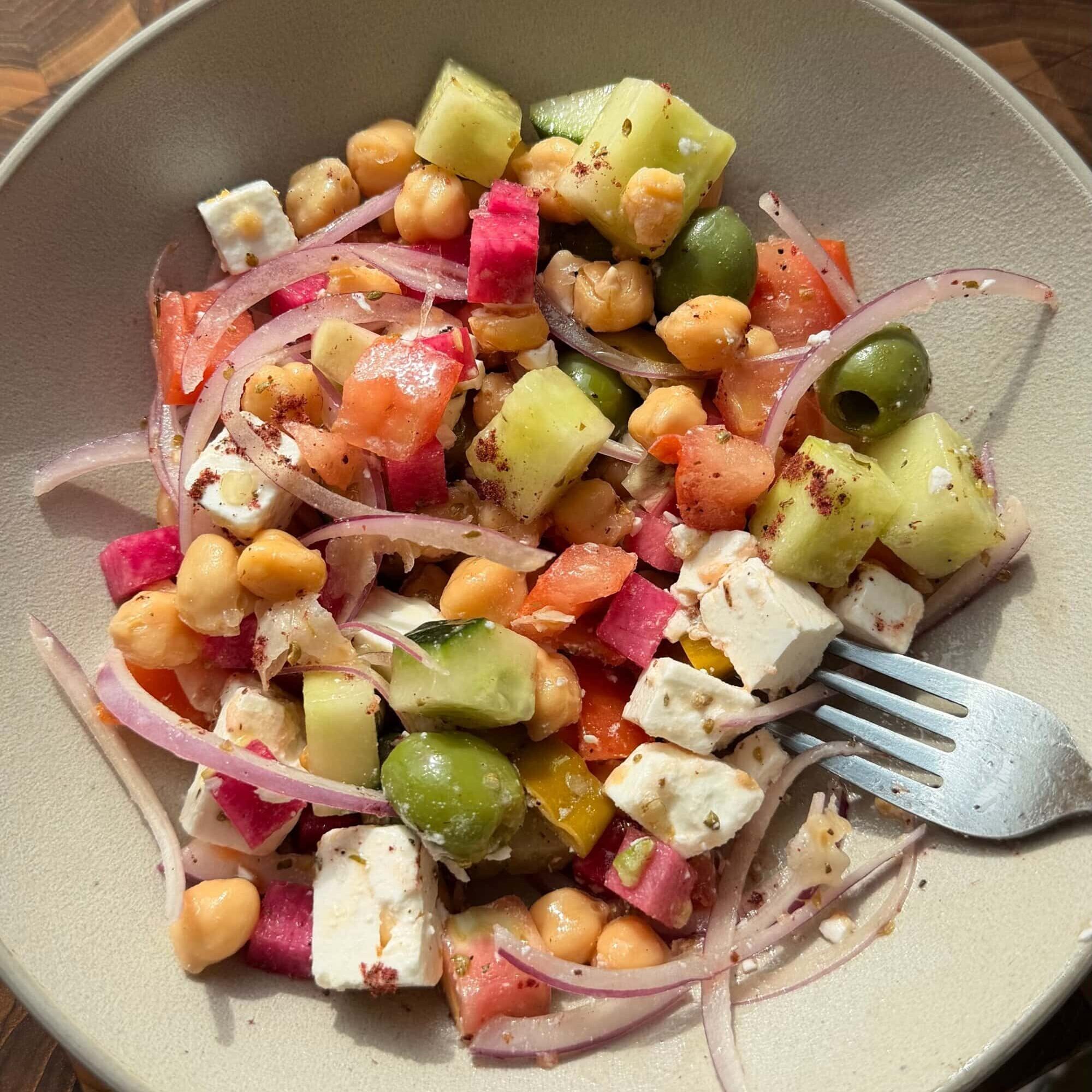 A bowl of salad with chickpeas, feta cheese, cucumber, green olives, diced tomatoes, red onion, and watermelon radish, with a fork on the side.