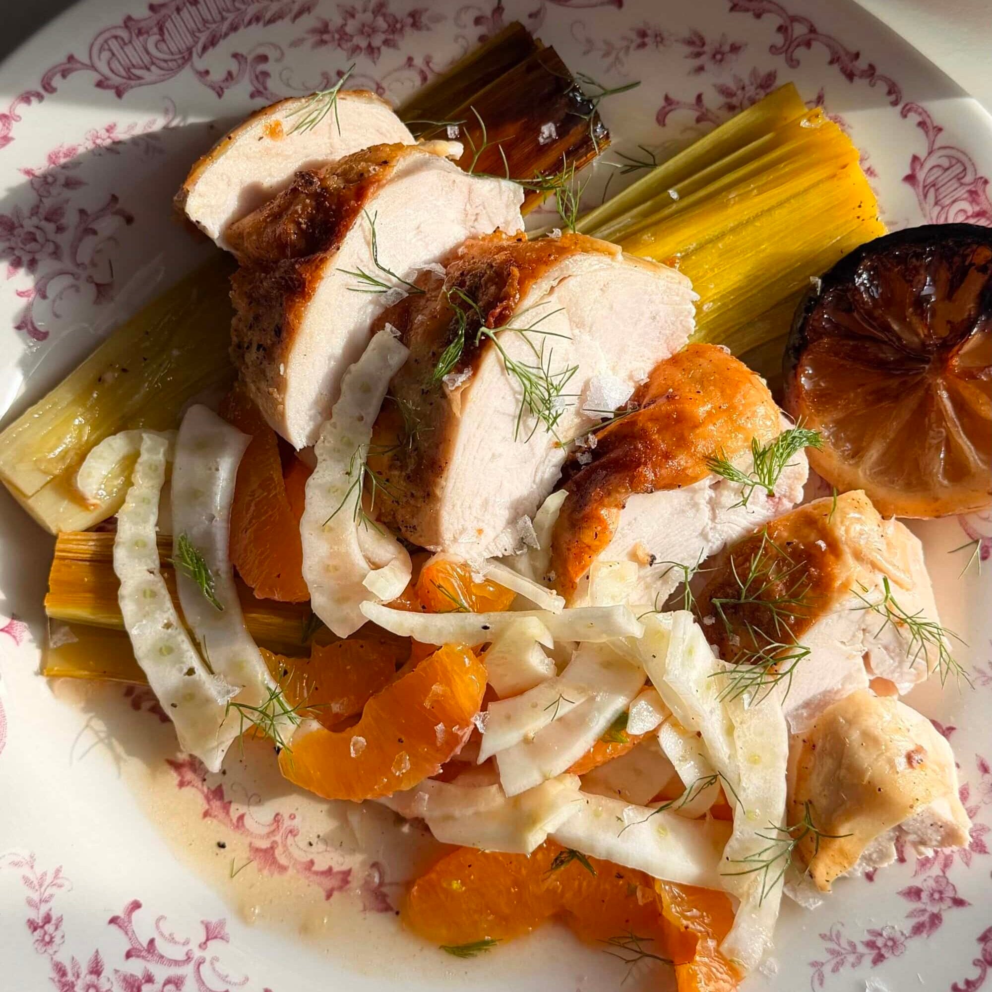 Sliced roasted chicken breast on a fennel and orange salad with braised leeks and a charred lemon half, served on a white floral-patterned plate.