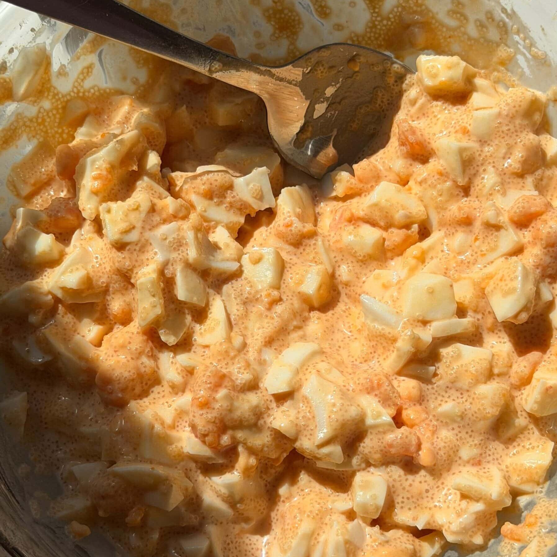 A glass bowl containing chopped hard-boiled eggs mixed with an orange-tinted creamy dressing, being stirred with a metal spoon.