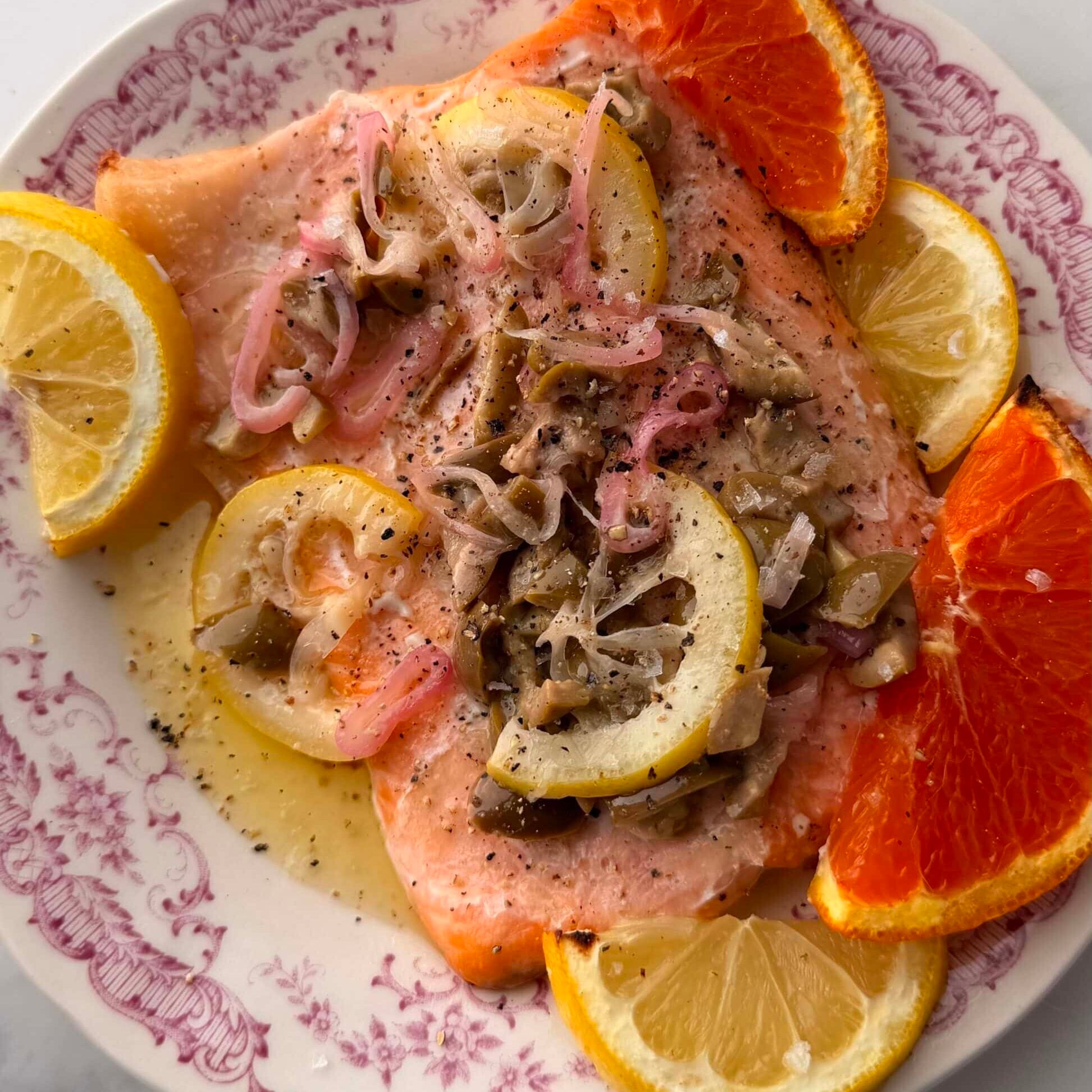 A plate with baked salmon topped with onions and mushrooms, garnished with lemon and orange slices on a white and pink patterned dish.