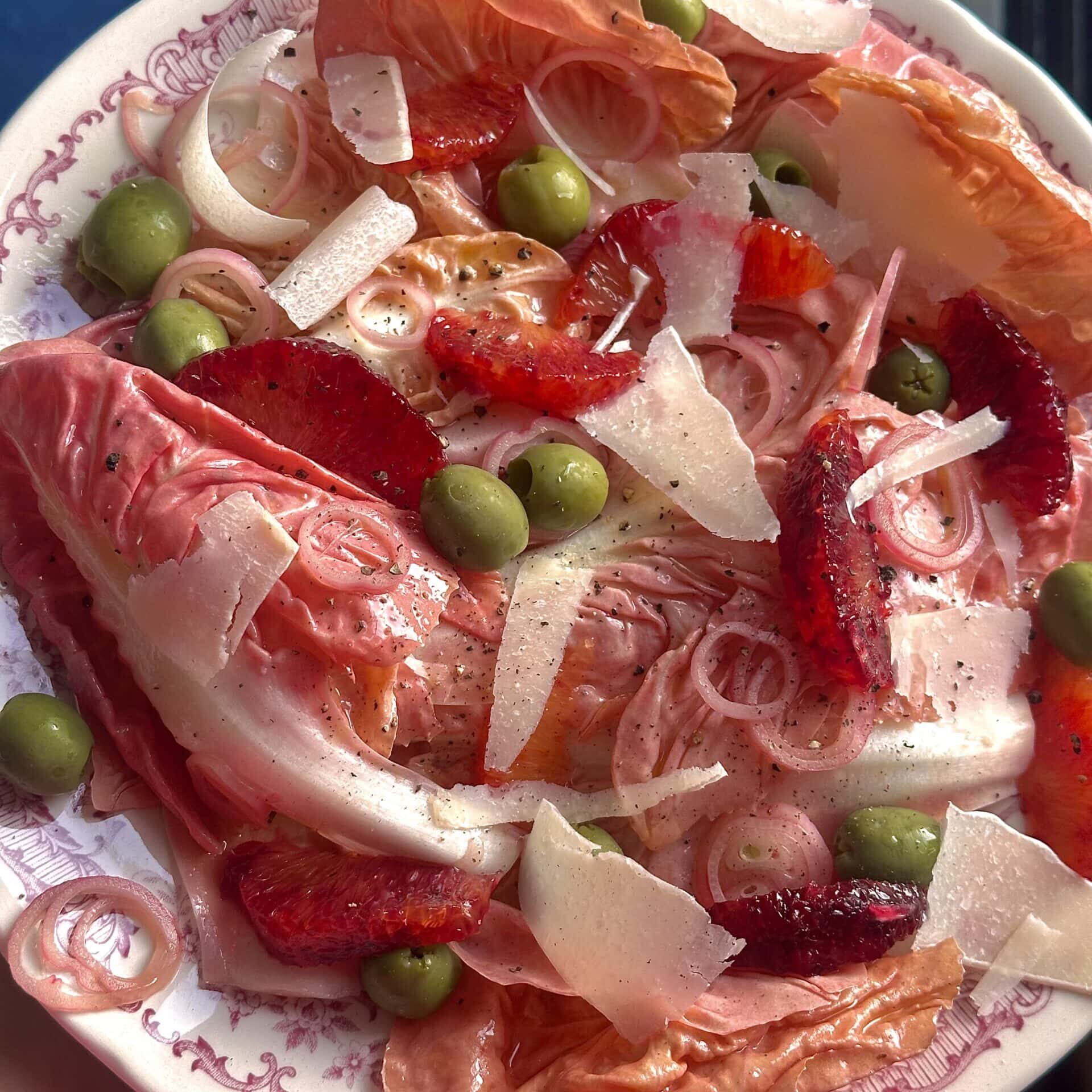 A plate of salad with red lettuce, green olives, sliced strawberries, shallots, and shaved cheese on a patterned dish.