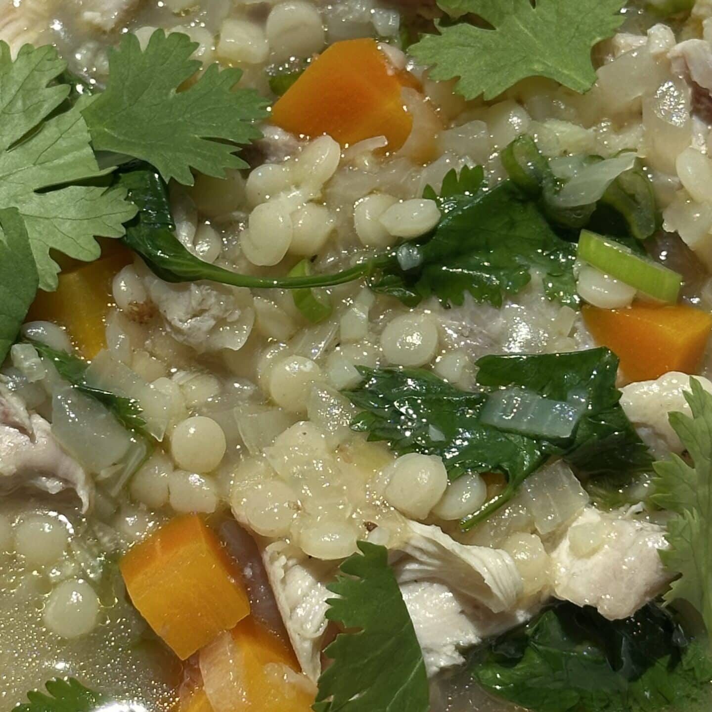 Close-up of a soup with diced carrots, shredded chicken, small pasta, leafy greens, and fresh cilantro in a clear broth.