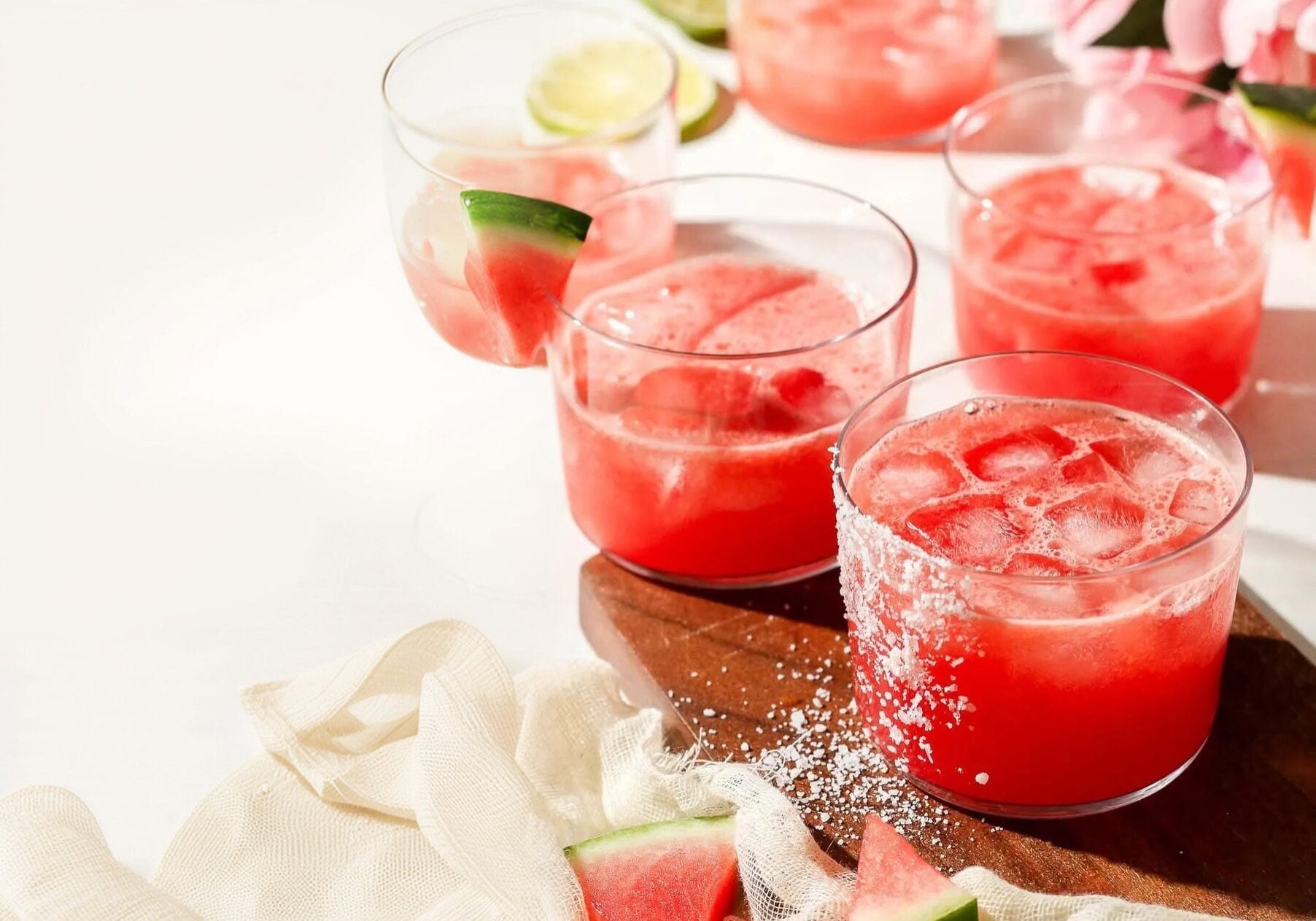 Five glasses of pink watermelon drinks with ice and watermelon wedges, some with salted rims, placed on a wooden board with lime and watermelon slices nearby.