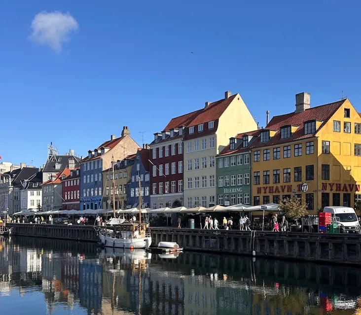 Colorful buildings line the Nyhavn waterfront in Copenhagen, Denmark, with boats docked along the canal under a clear blue sky.