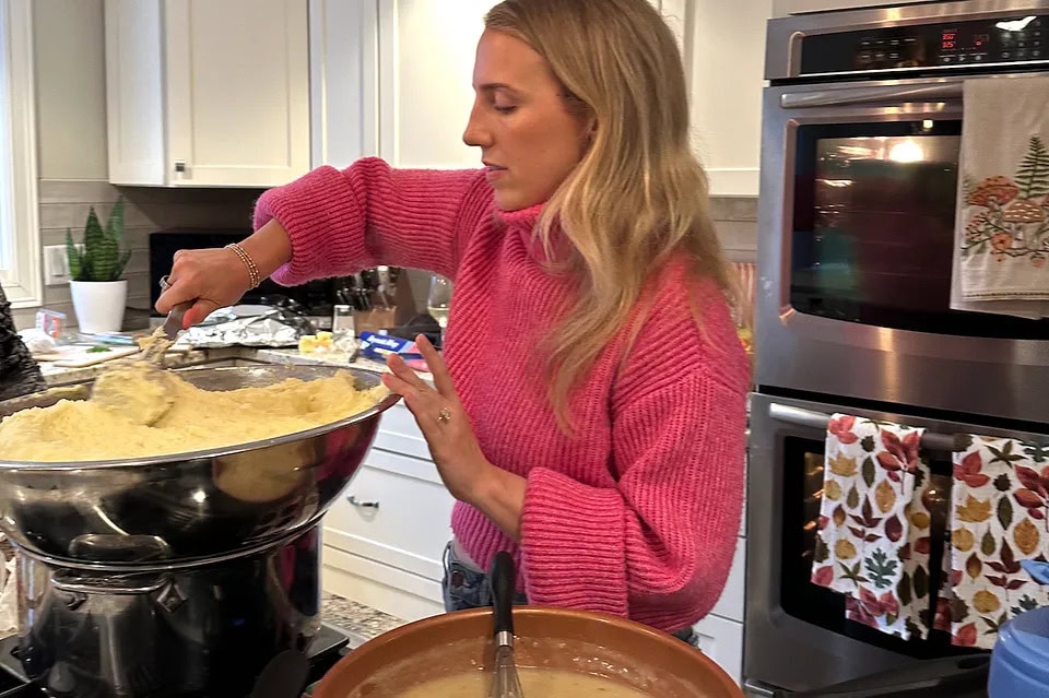 A woman in a pink sweater stirs a large bowl of batter in a kitchen, with an oven and mixing bowls visible in the background.
