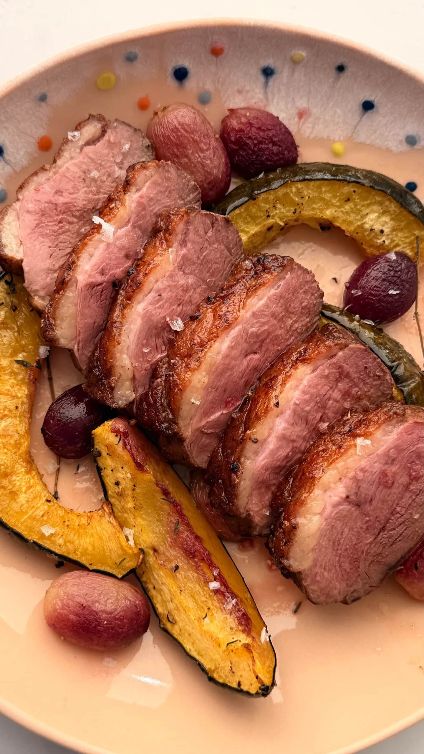 Sliced roasted duck breast served with roasted acorn squash wedges and red grapes on a polka-dotted plate.