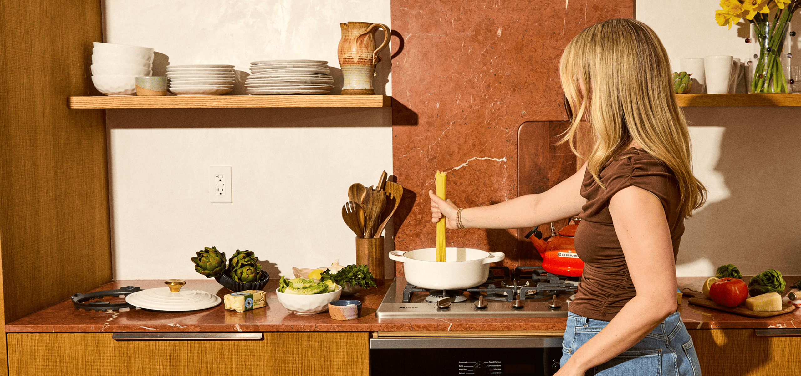 A woman with long blonde hair stirs a pot on a stove in a kitchen with wooden cabinets, plates, vegetables, and cooking utensils on the counter.