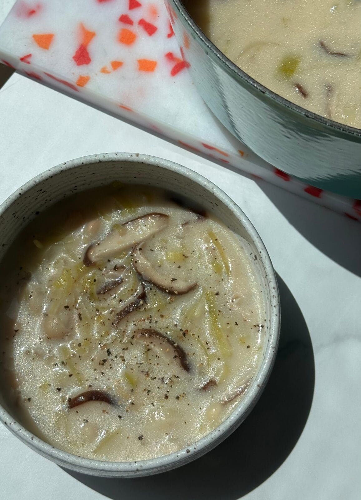 A bowl of creamy mushroom soup with visible mushroom slices and ground pepper, next to a pot containing more of the same soup.