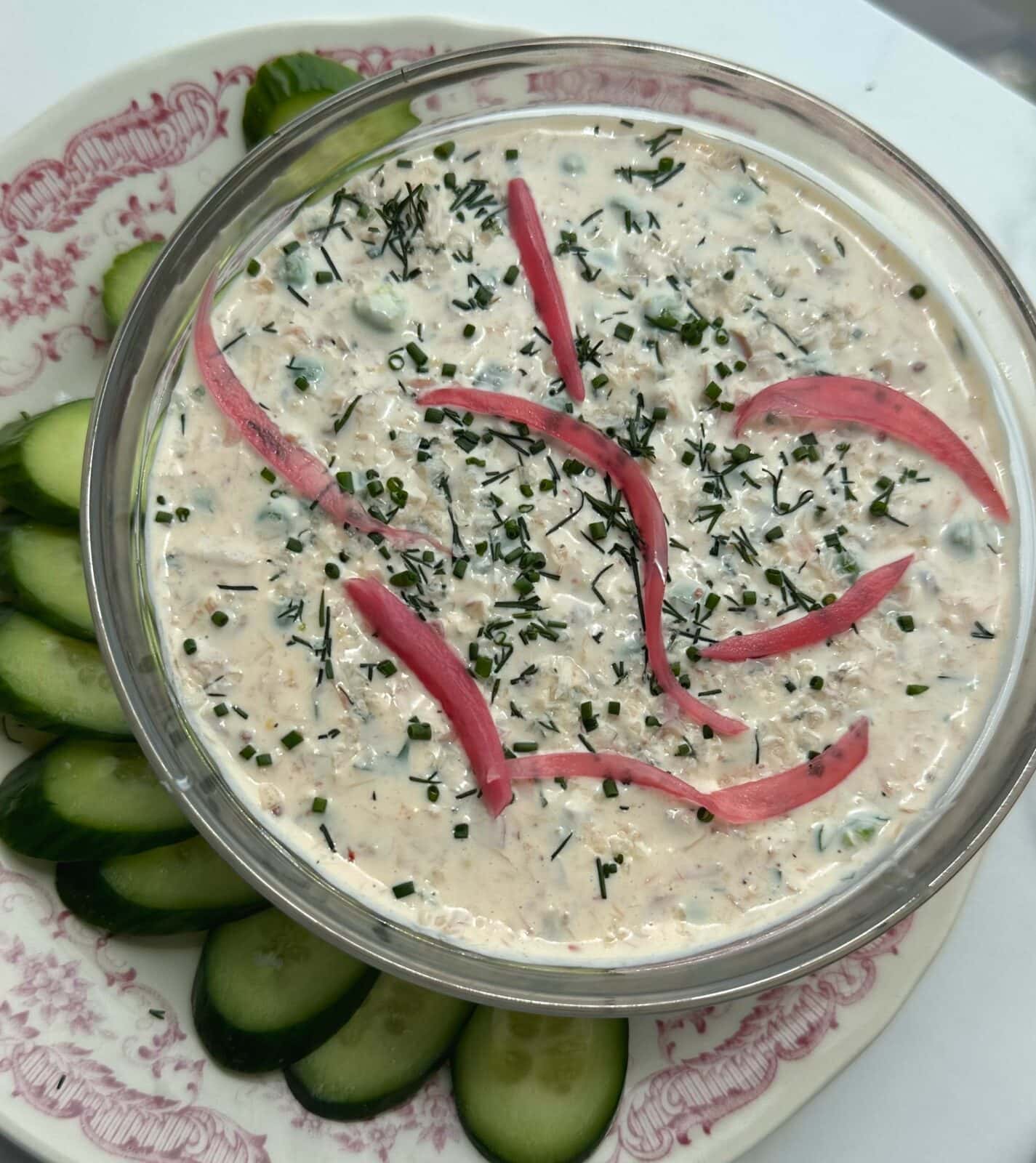 A bowl of creamy dip garnished with chopped herbs and pickled onions, surrounded by sliced cucumbers on a decorative plate.