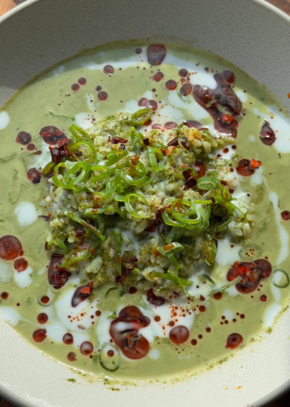 A bowl of green soup topped with a dollop of rice, sliced scallions, white cream, and red chili oil drizzles.