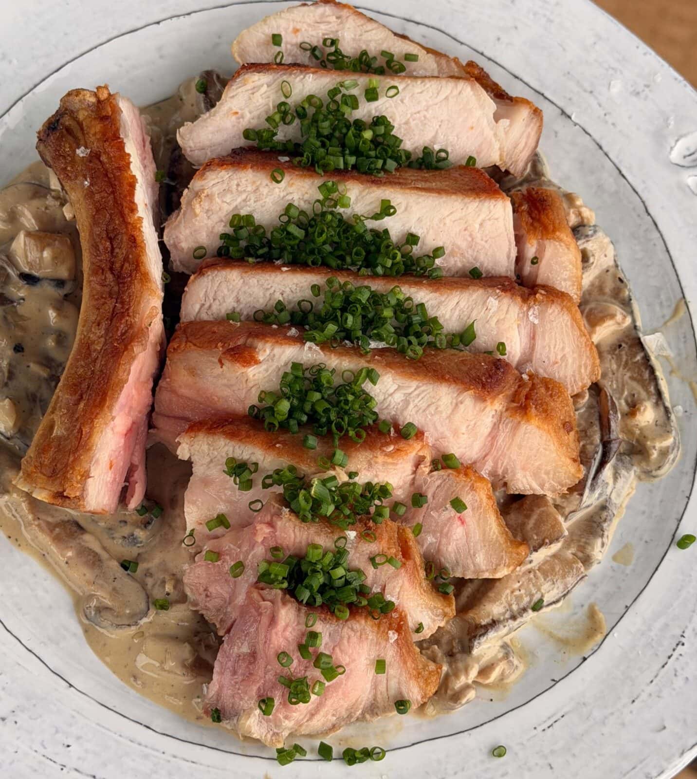 Sliced pork with a crispy edge served over creamy mushrooms, garnished with chopped chives on a white plate.