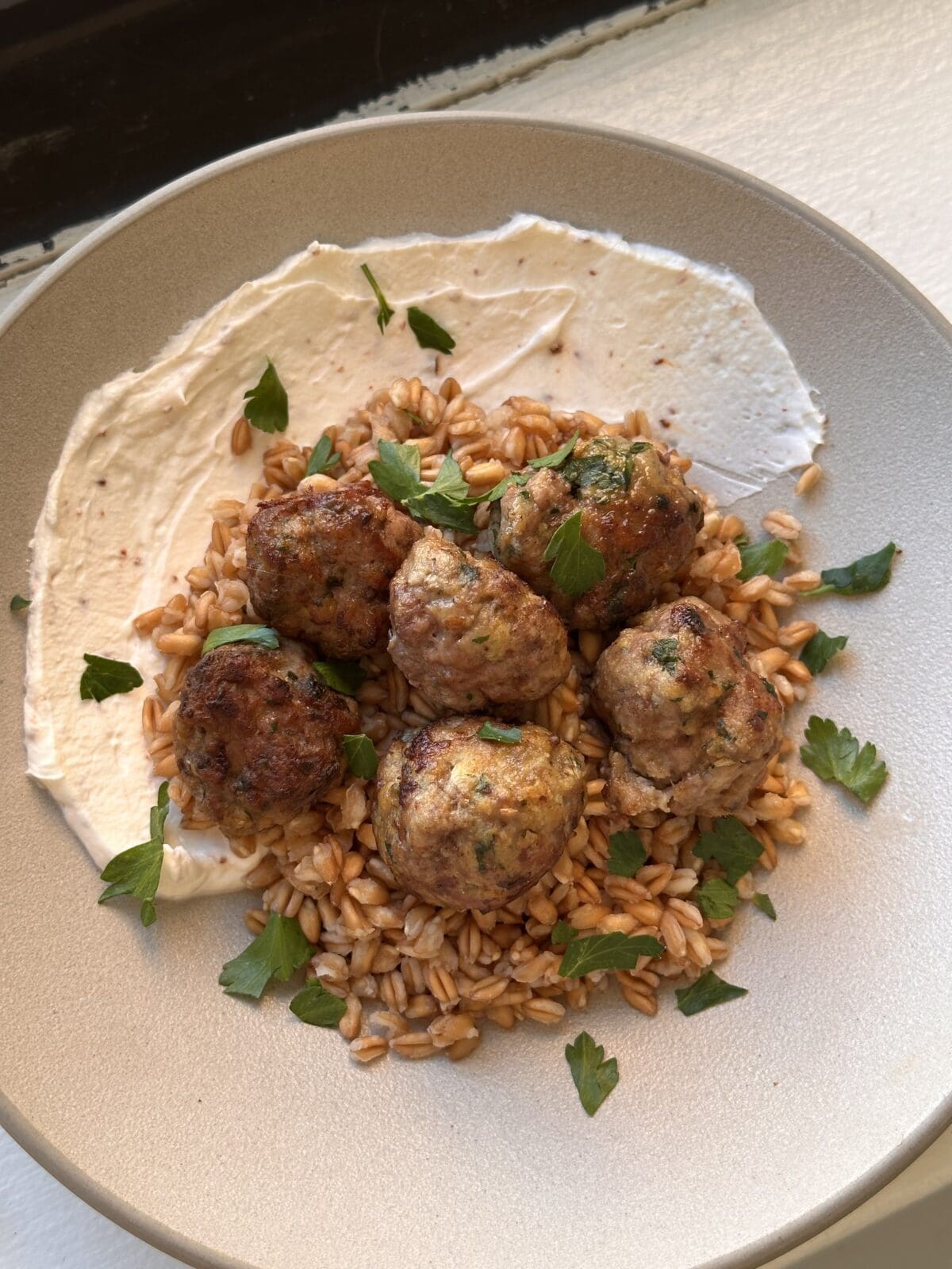 Five meatballs on a bed of grains, served with a smear of white sauce and garnished with fresh parsley on a beige plate.