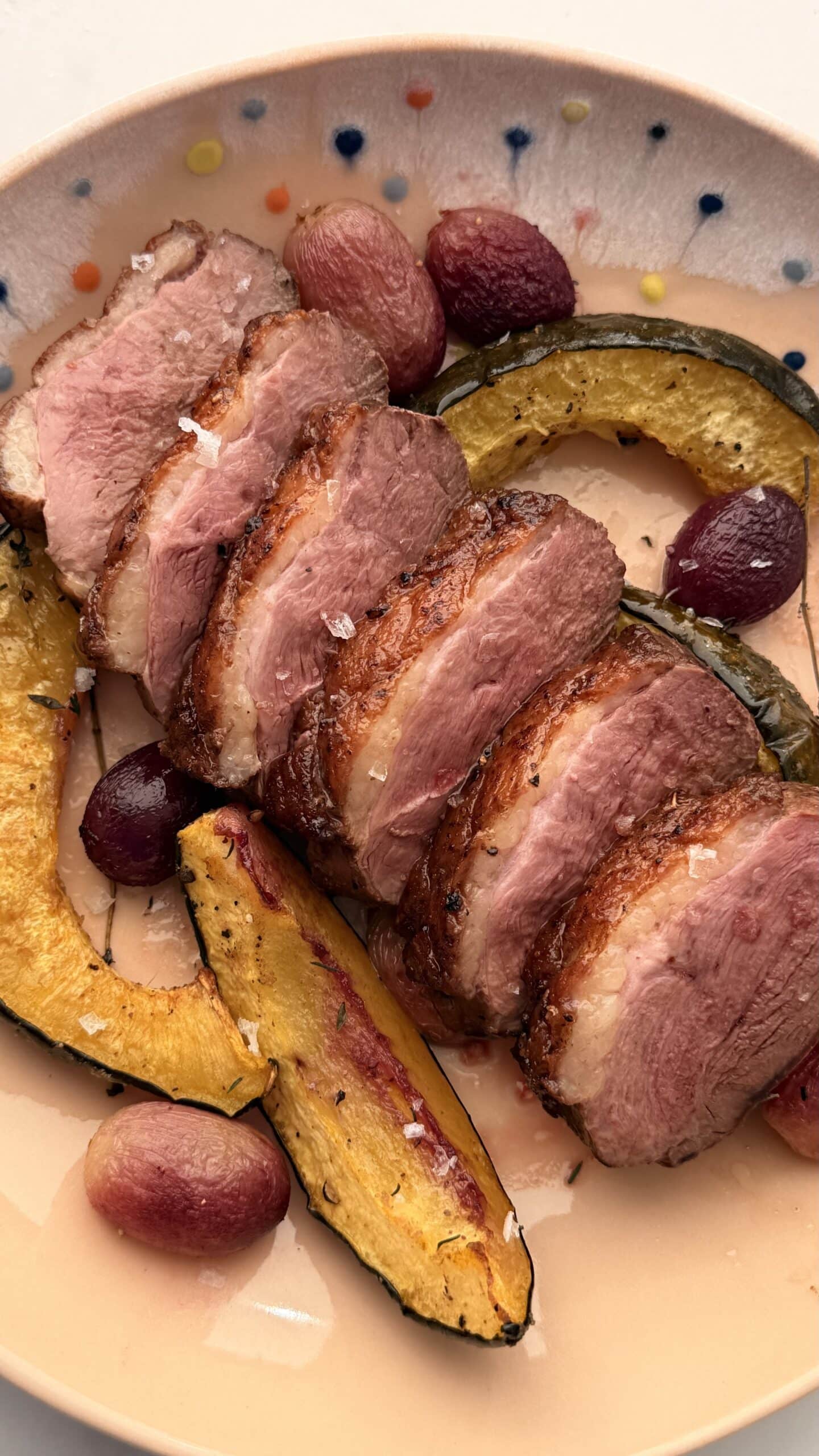 Sliced roasted duck breast served with roasted squash and red grapes on a speckled ceramic plate.