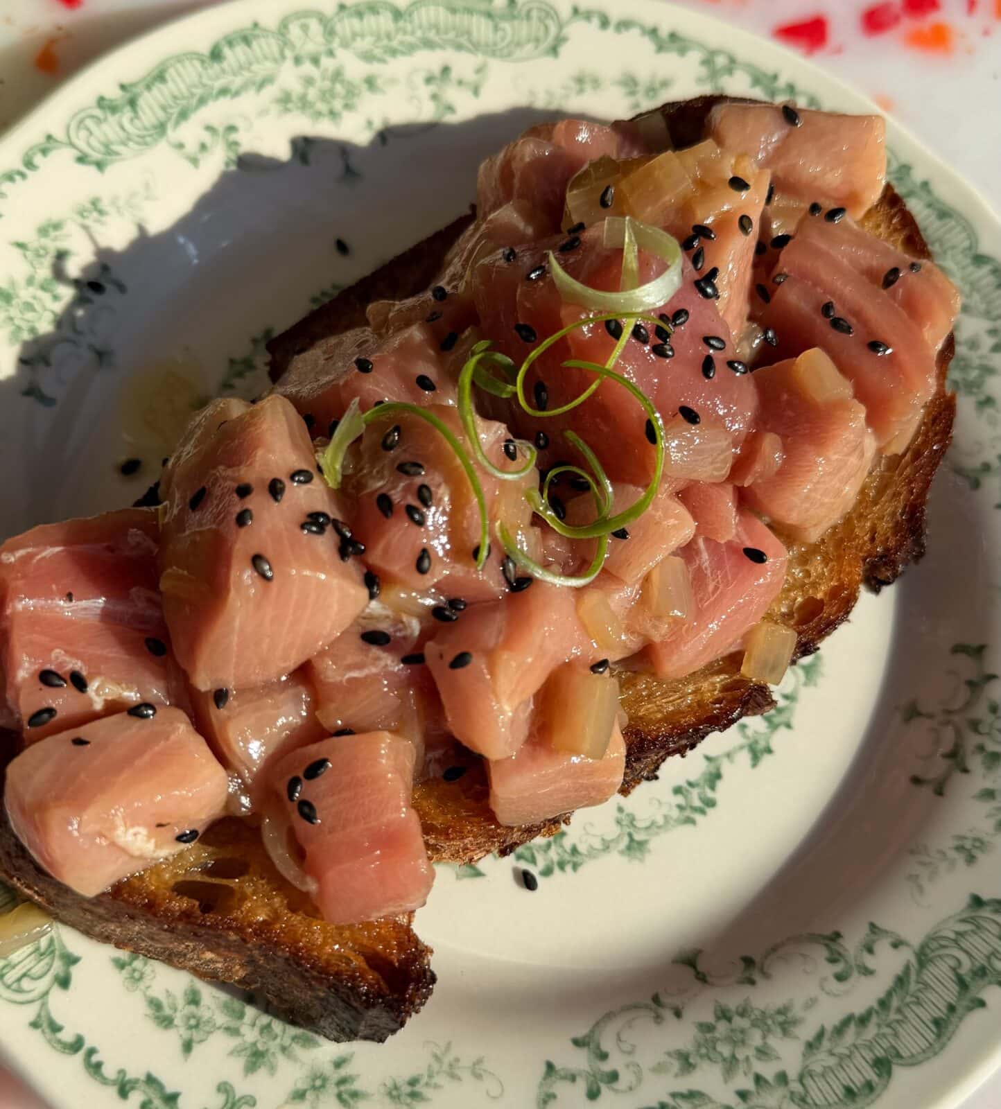 A slice of toasted bread topped with diced raw fish, garnished with black sesame seeds and thinly sliced green onions, served on a decorative plate.