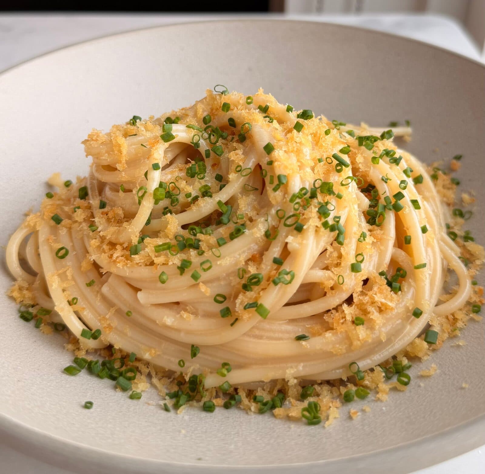 A plate of spaghetti topped with finely chopped chives and golden breadcrumbs, served in a shallow white bowl.