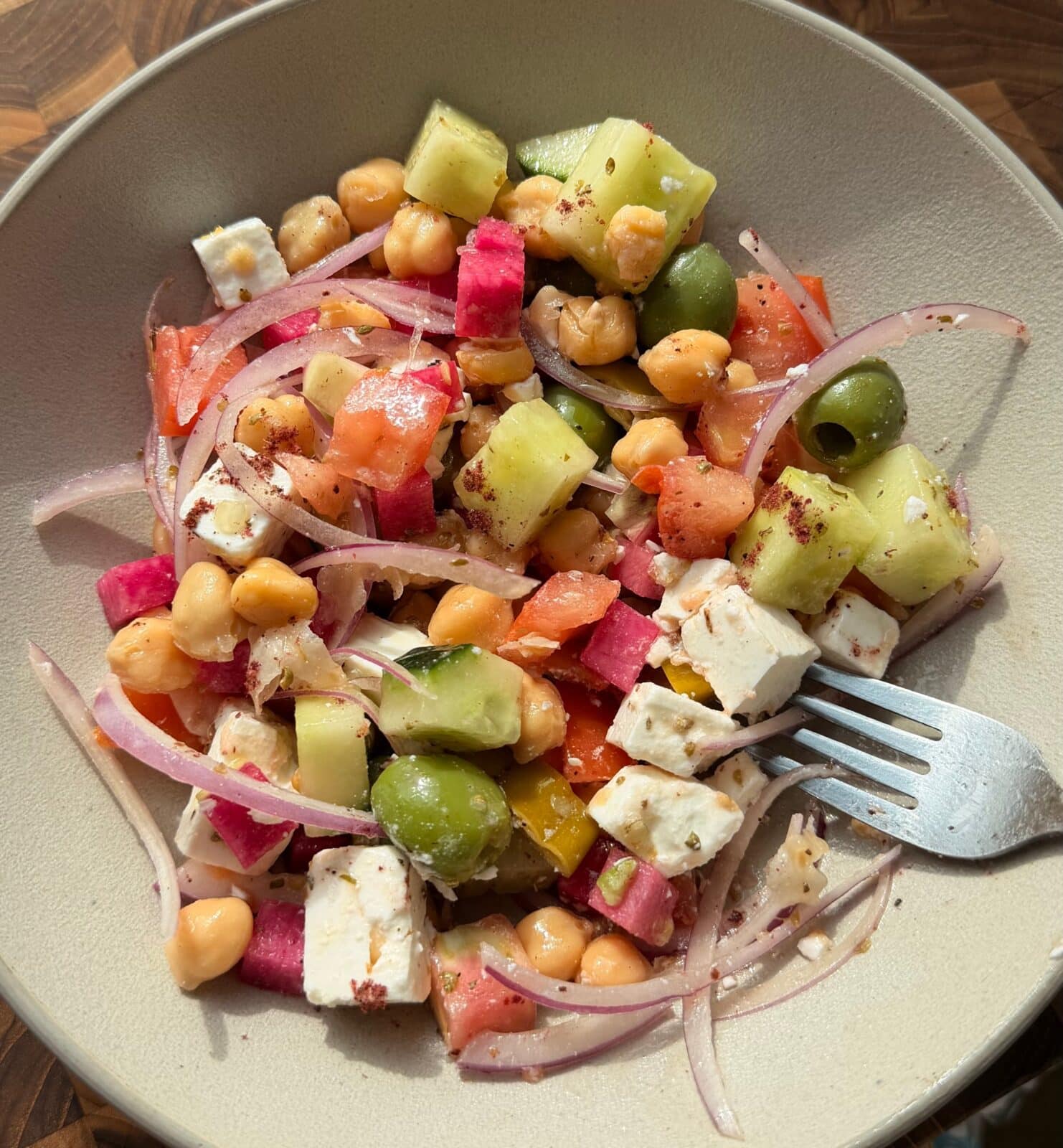 A bowl of salad with chickpeas, feta cheese, cucumber, green olives, diced tomatoes, red onion, and watermelon radish, with a fork on the side.